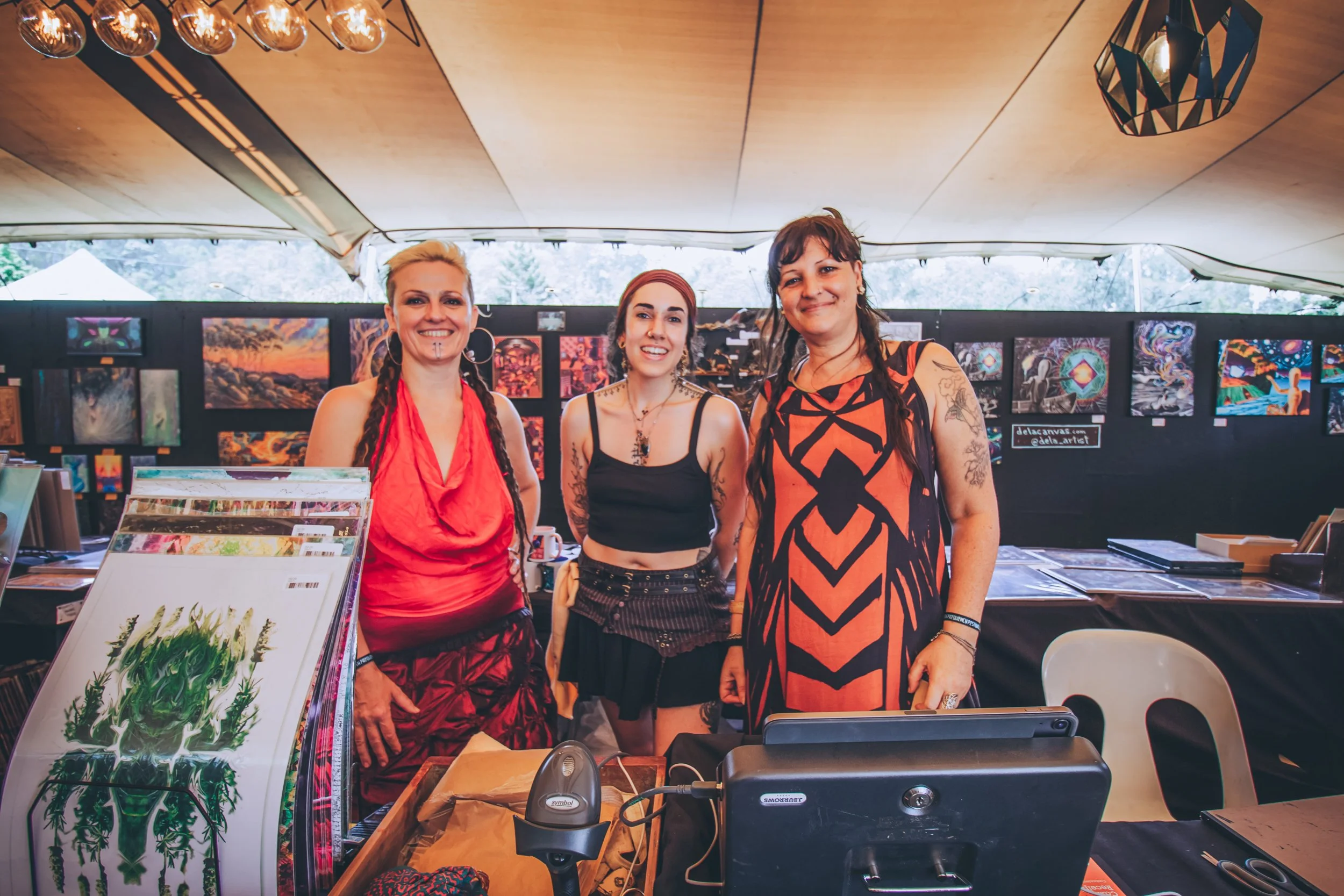Three women standing behind a booth at an art fair with paintings displayed on the back wall and art prints on the table in front of them.