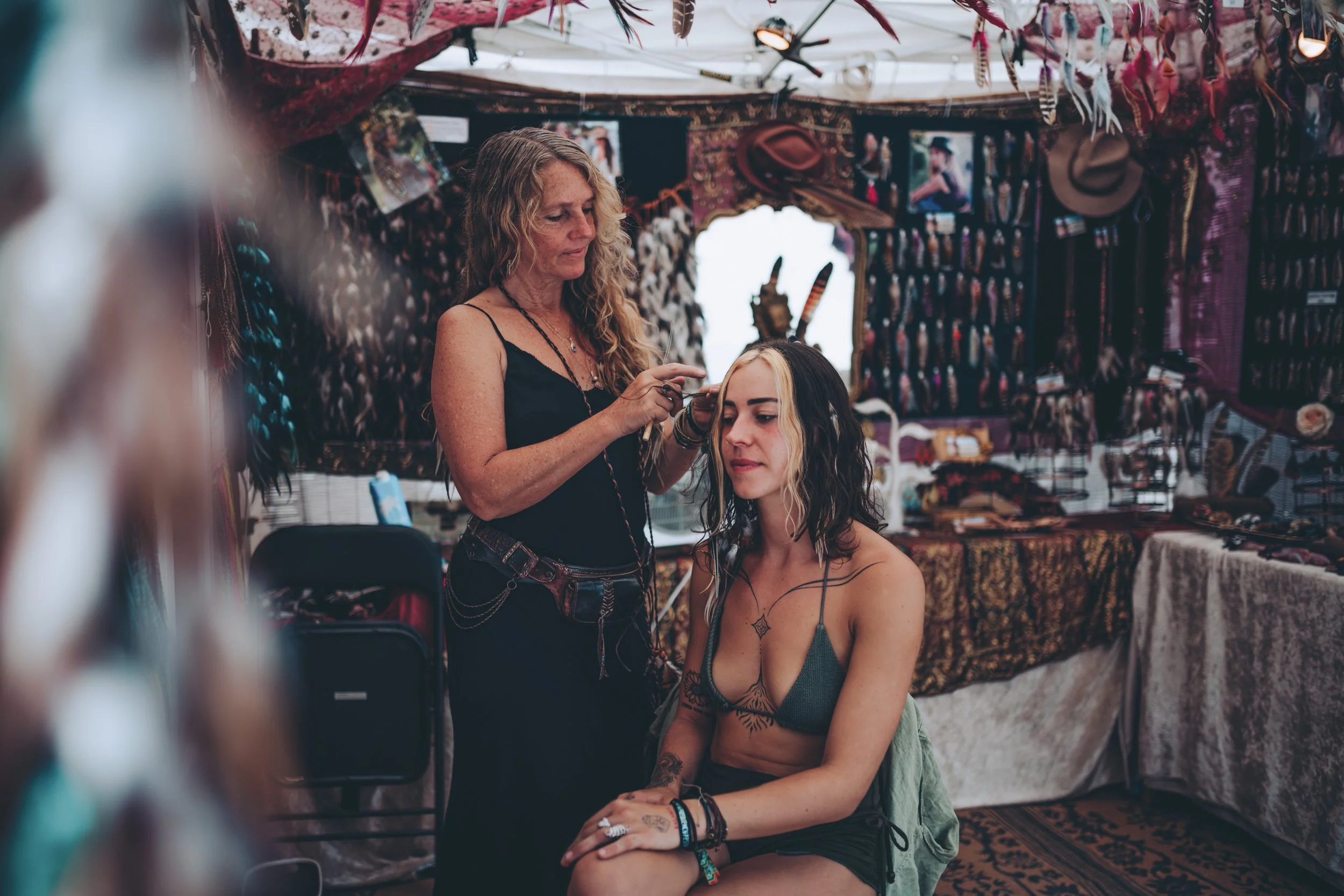 A woman is sitting with her eyes closed while a female artist applies makeup or face paint in a decorated booth, with photographs, hats, and jewelry displayed behind them.