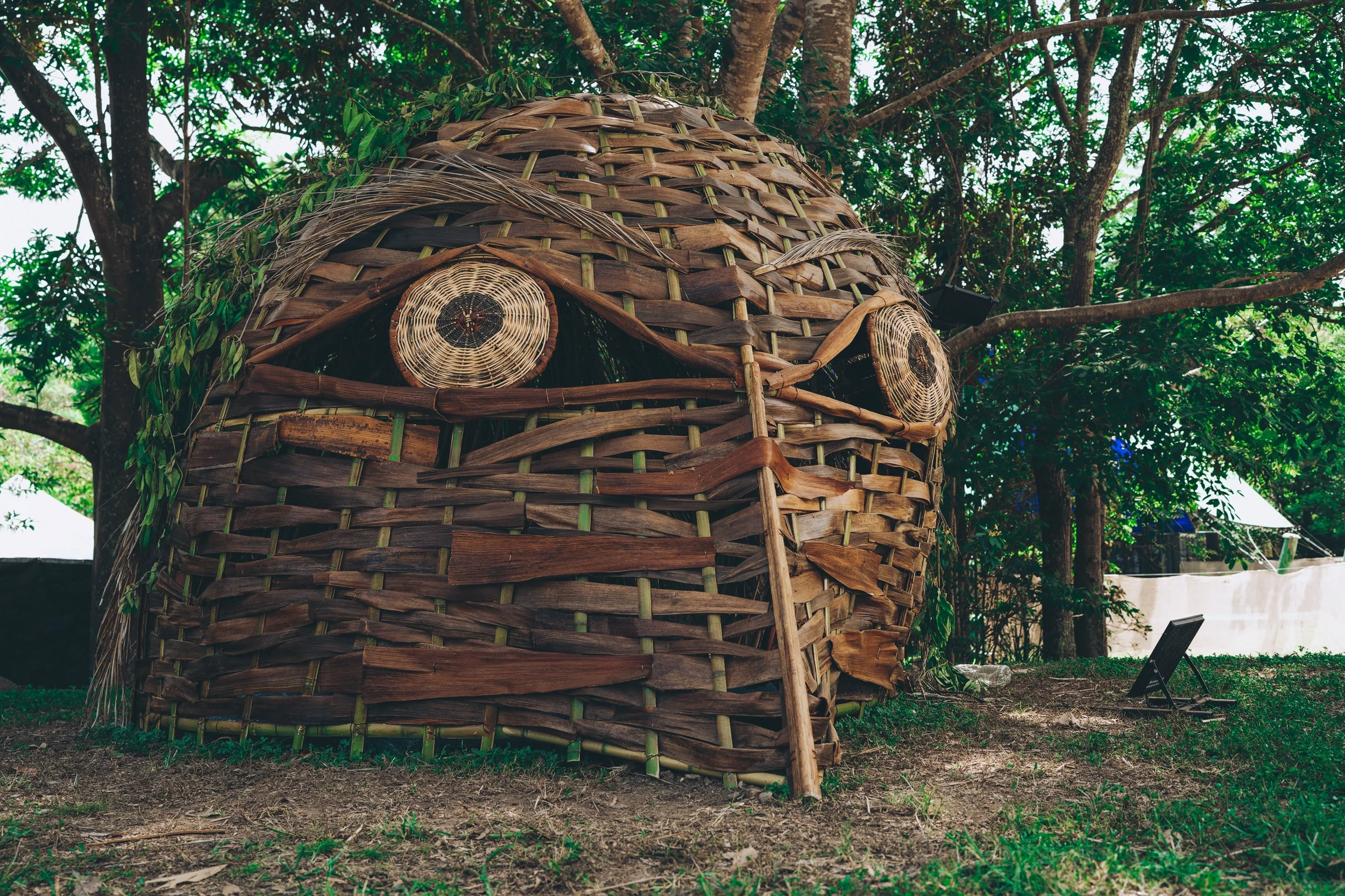A large, woven bamboo structure resembling an owl, with circular woven details for eyes, is situated outdoors among trees and grass.