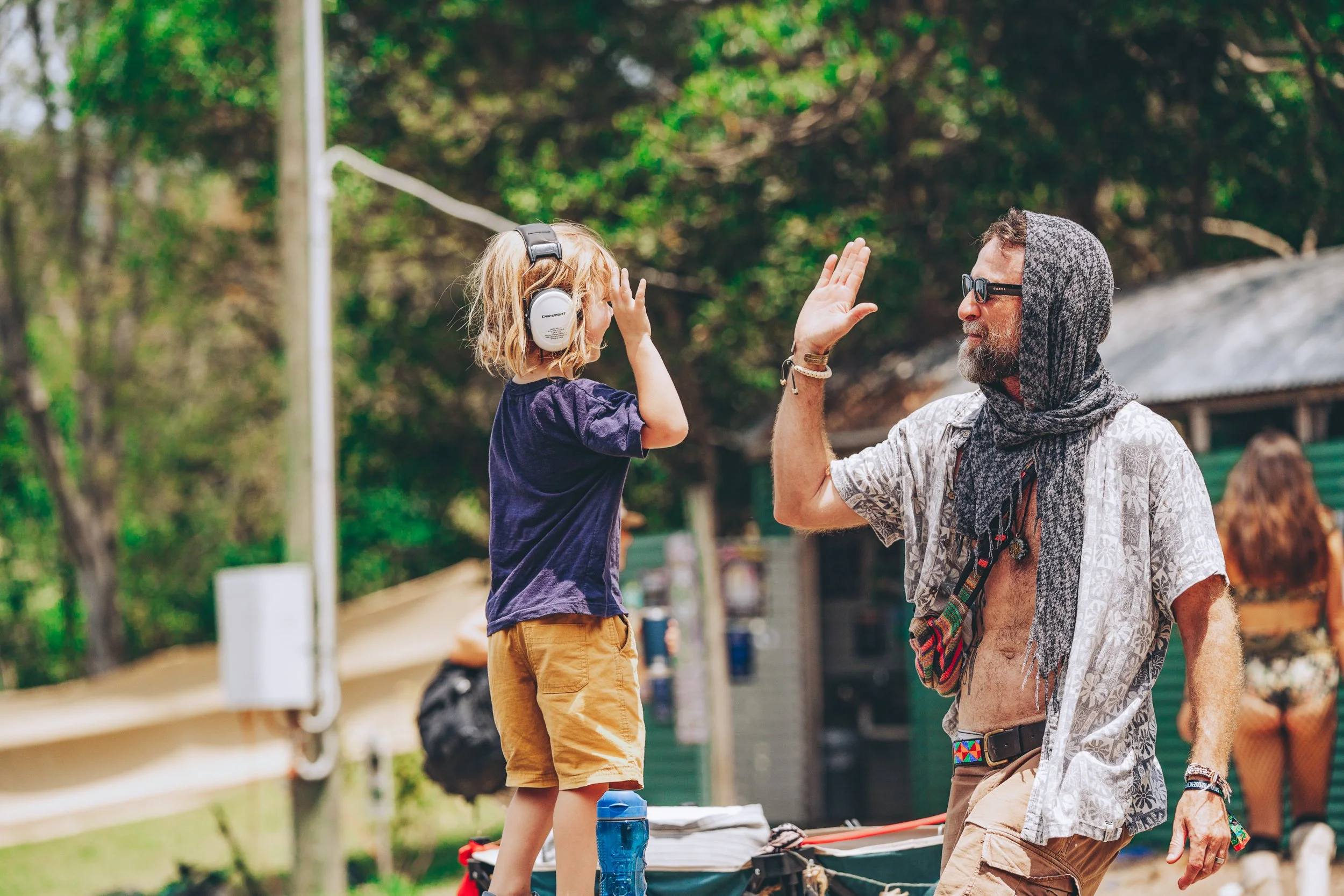 A man and a young boy engaging in a high five outdoors in a wooded area. The boy is wearing headphones and a dark T-shirt, standing on a surface with a water bottle and other items nearby. The man is wearing sunglasses, a hooded scarf, and a casual s