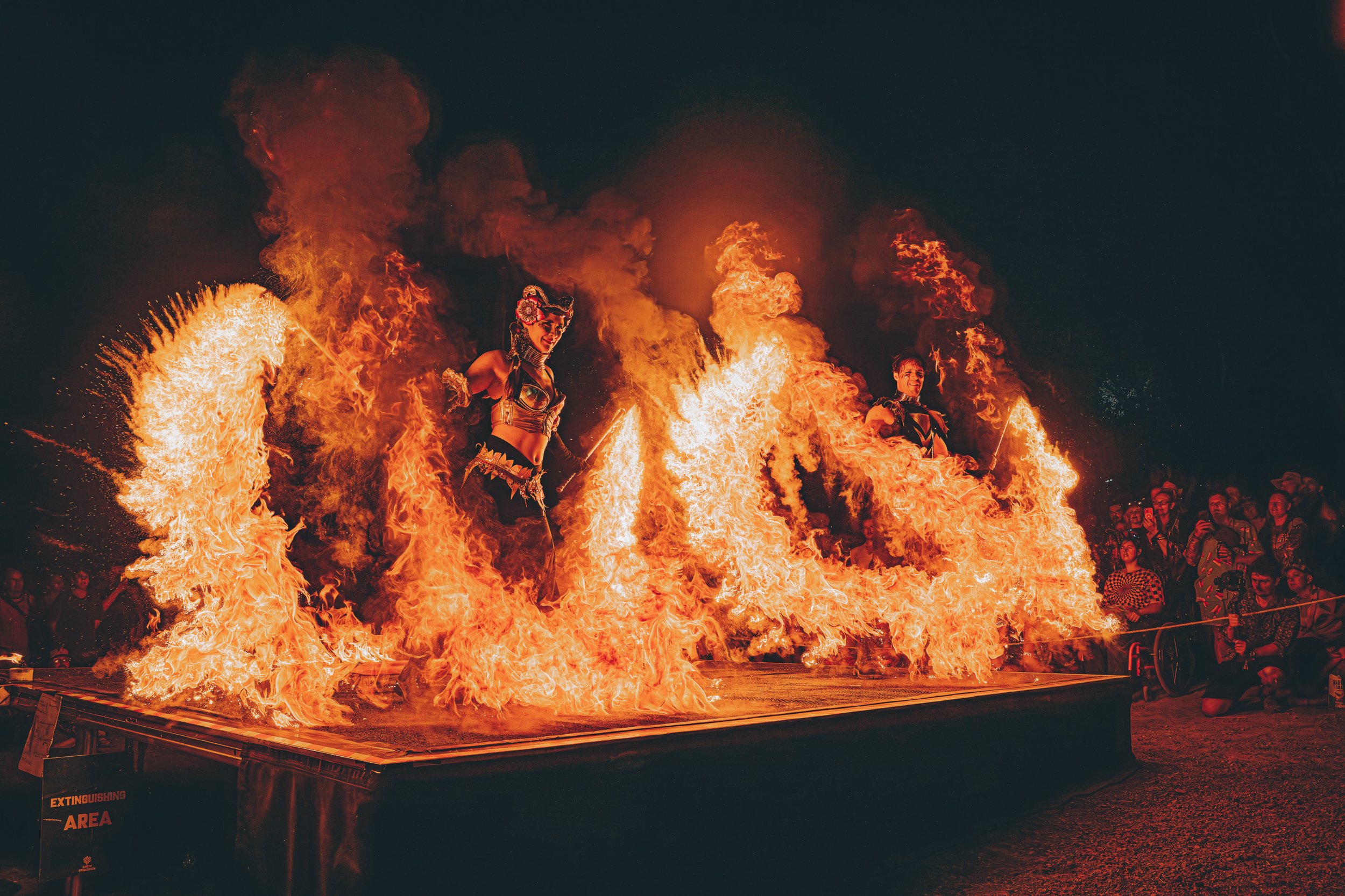 Fire dancers performing on stage with flames surrounding them, and audience watching at night.
