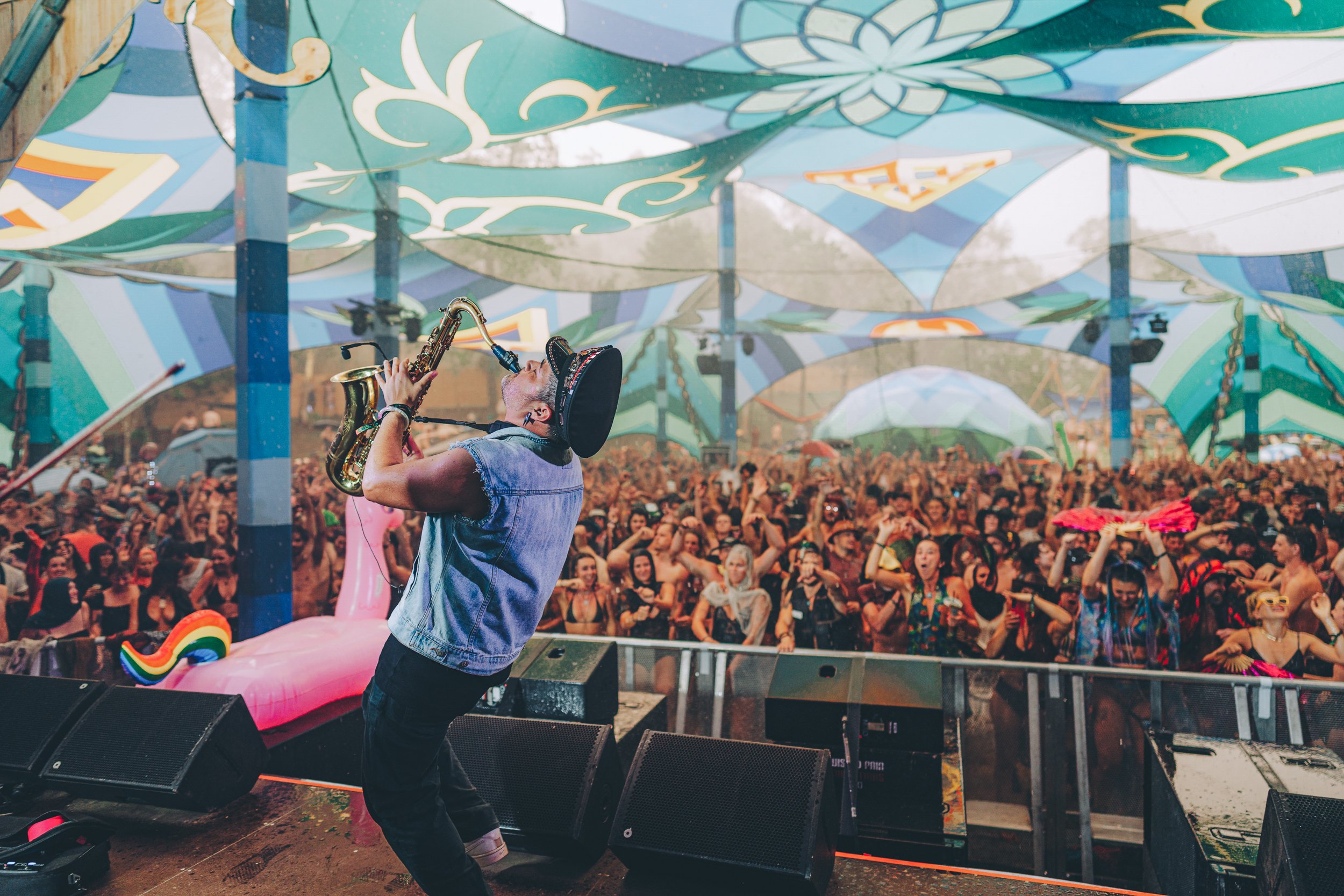 A DJ on stage playing a saxophone at an outdoor music festival with a large crowd dancing and enjoying the event.