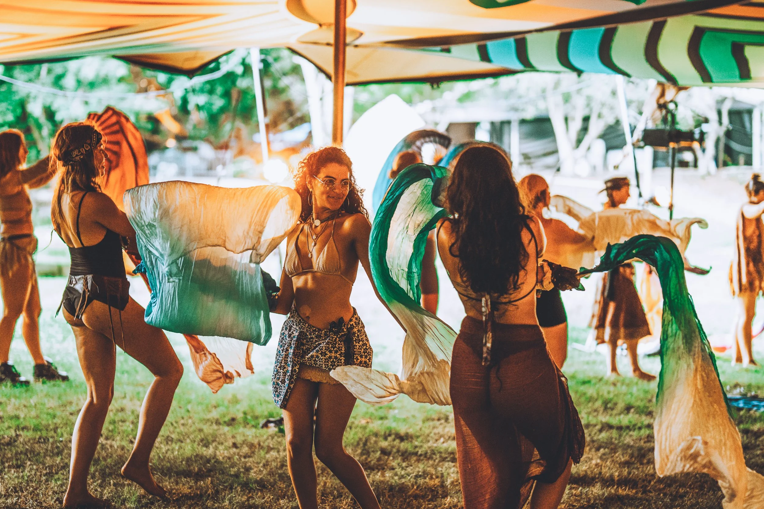 People dancing with large, colorful fabric pieces under a tent at an outdoor event during sunset.