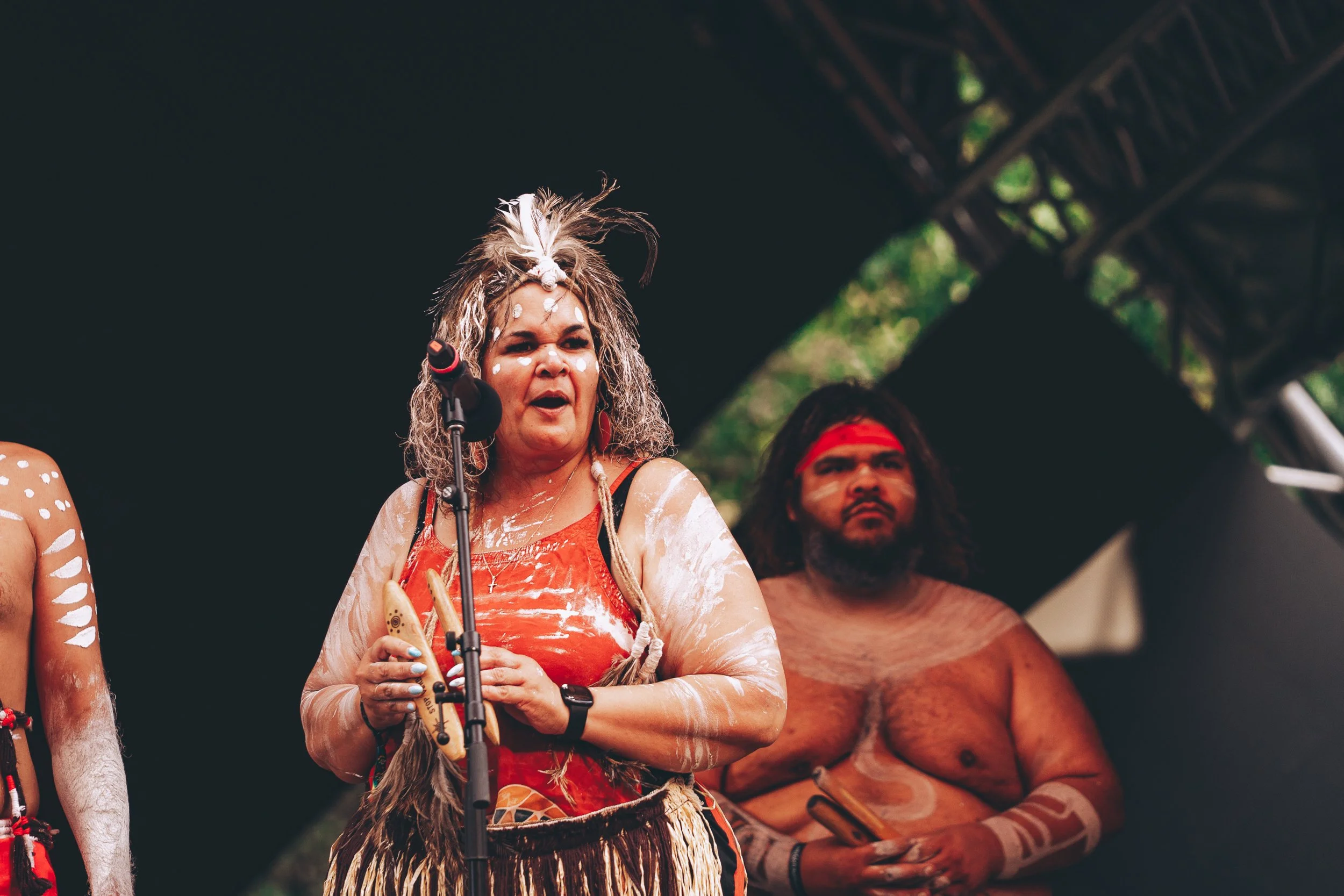 Indigenous people in traditional attire participating in a cultural event or ceremony, with one woman speaking into a microphone and a man standing behind her, under a shelter with trees visible in the background.