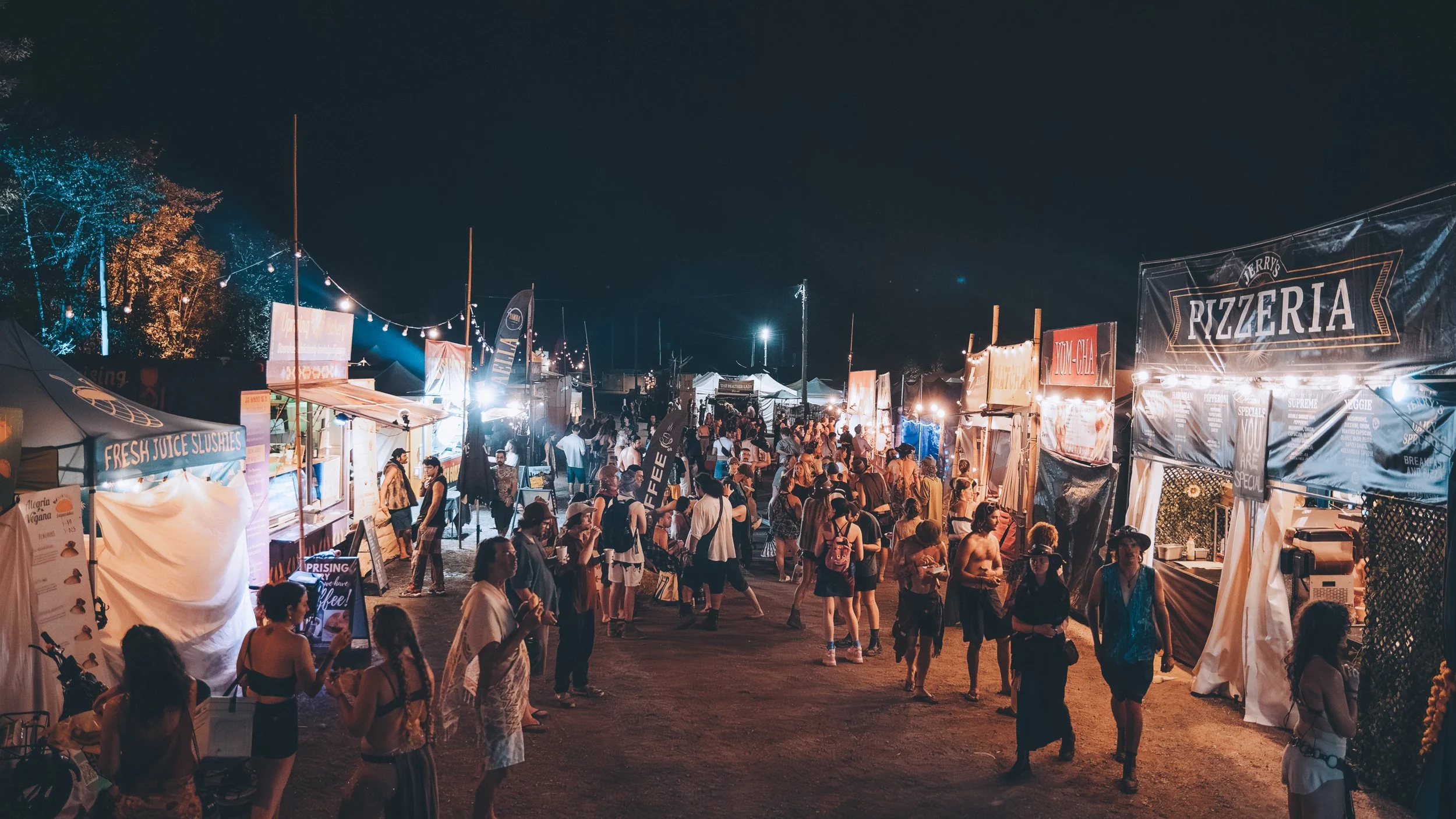 Night market with food stalls and string lights, crowded with people walking and shopping at outdoor festival.
