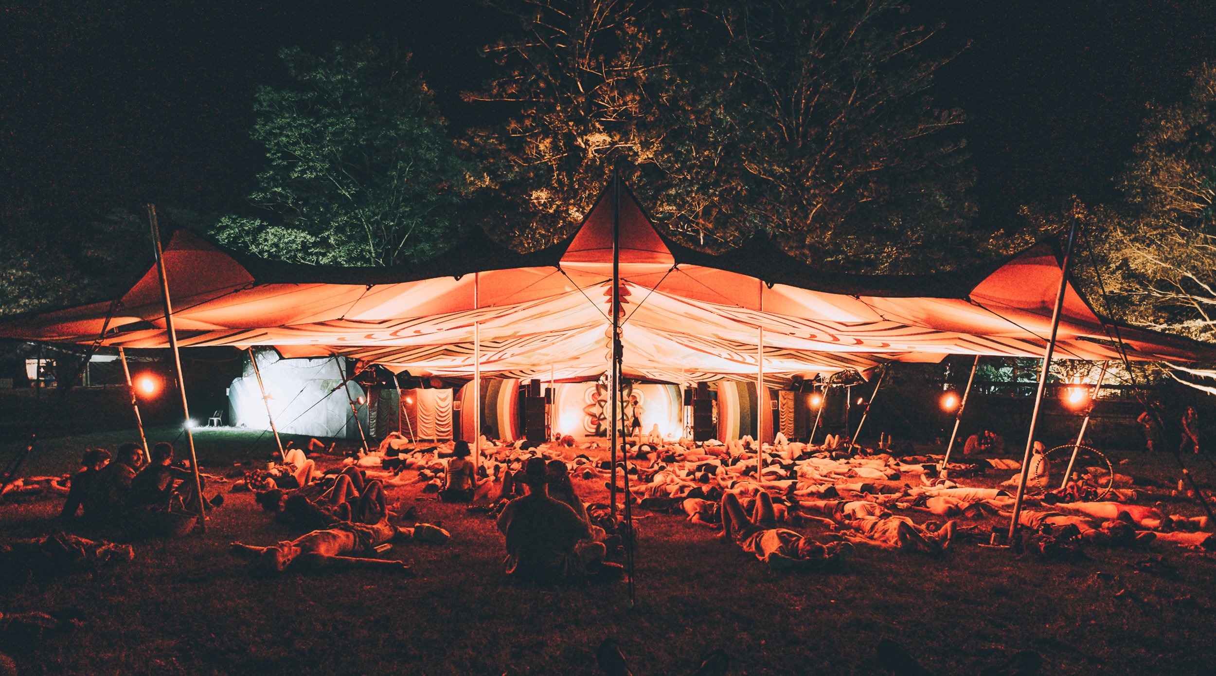 Outdoor night concert under large red and white canopy with stage, people lying on grass, and trees illuminated in the background.