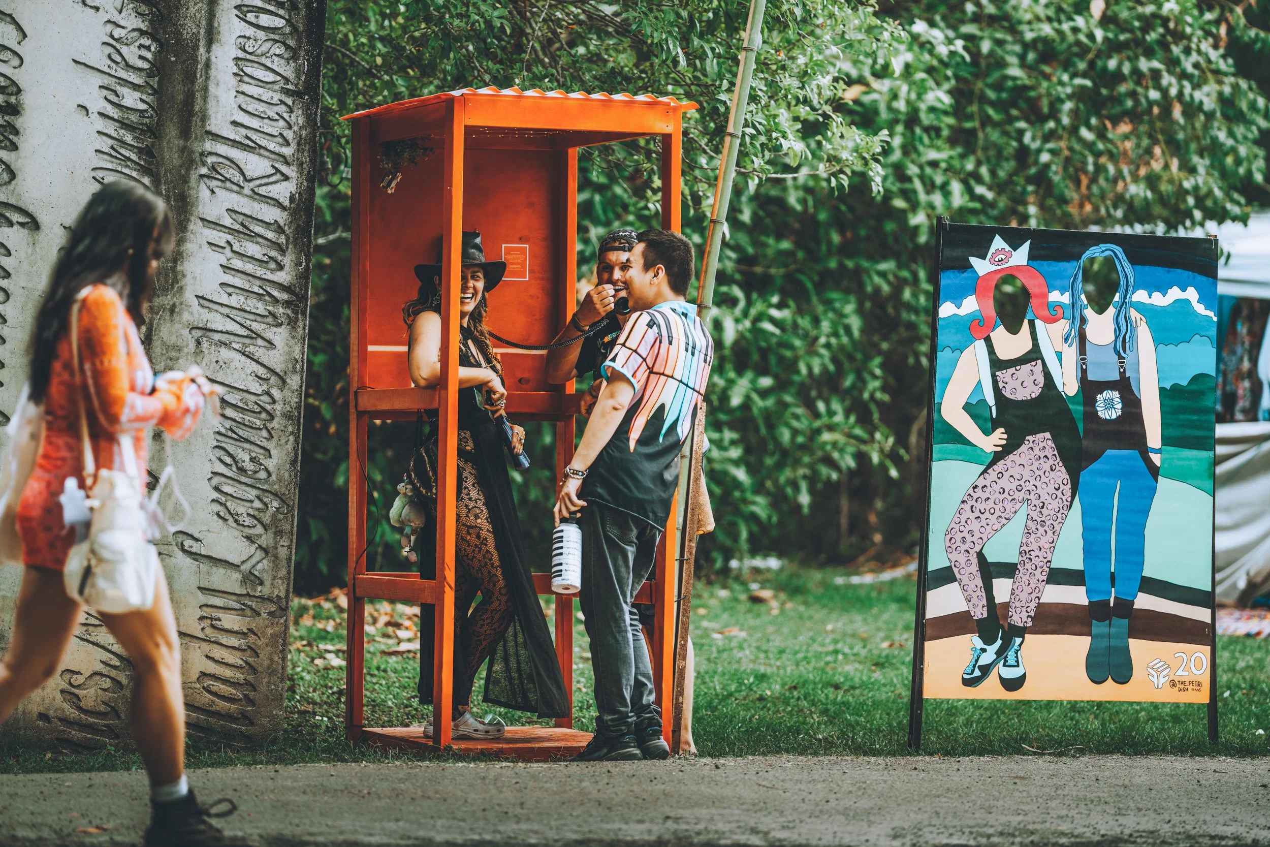 People interacting at an outdoor event with a red telephone booth and a colorful art display of two women in casual clothing, one with pink hair and the other with blue hair, against a background of trees and a large stone monument.