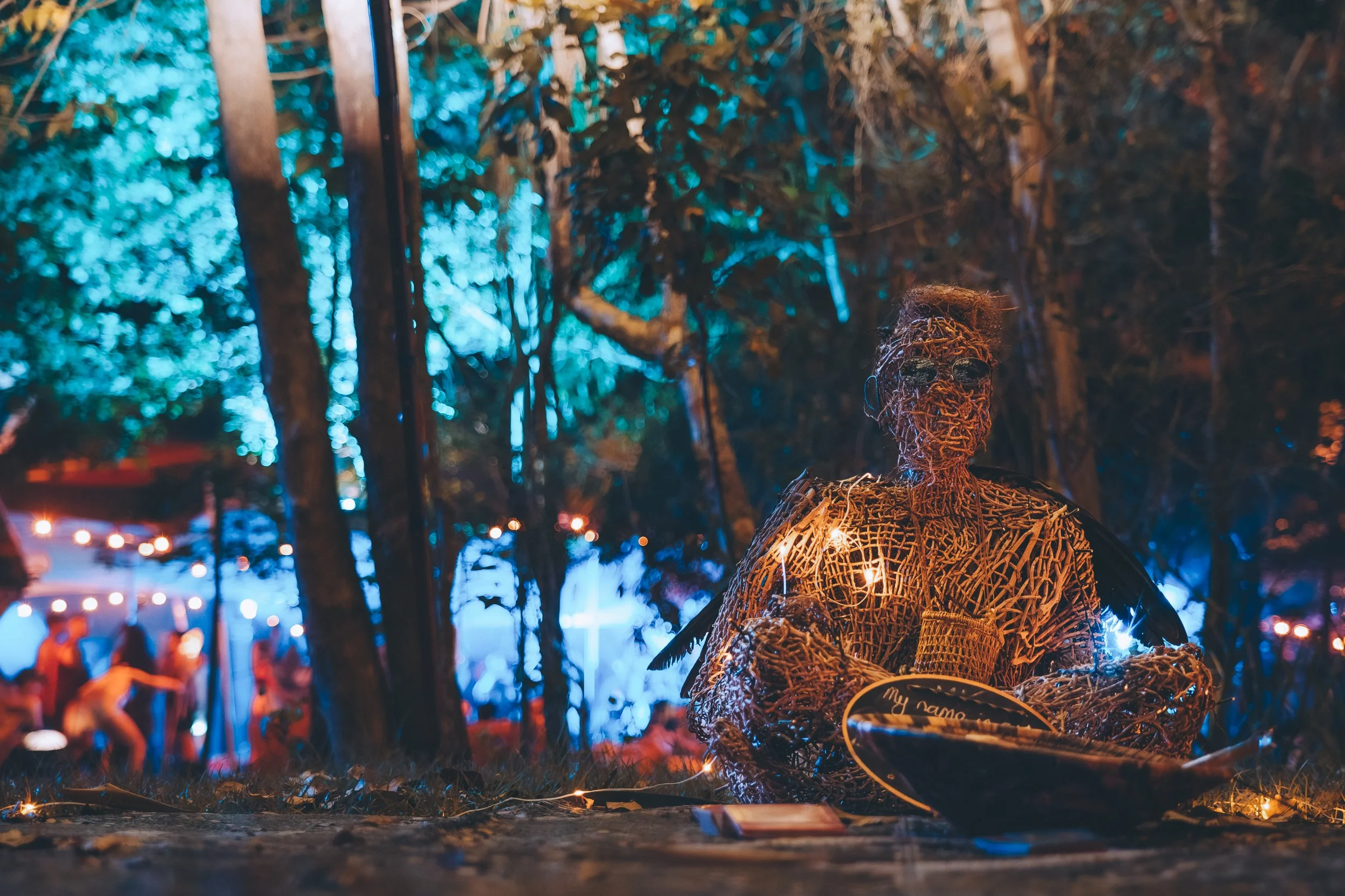 A woven art sculpture of a person with wings, holding a cup, illuminated by fairy lights, situated outdoors at night with blurred trees and colorful lights in the background.