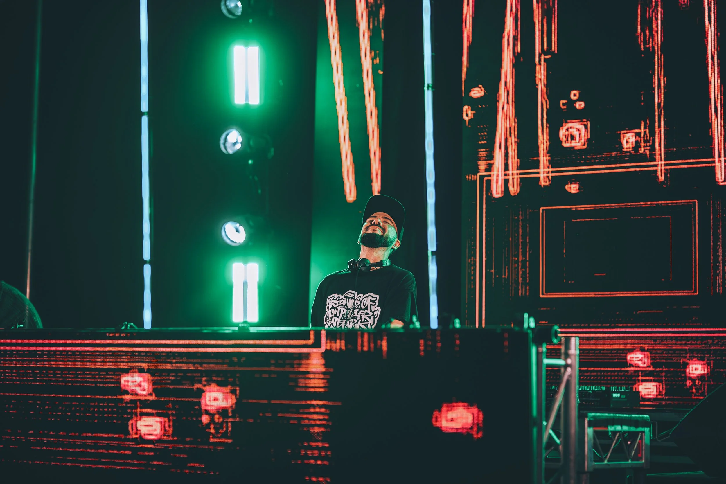 A DJ wearing a baseball cap and headphones stands behind a DJ booth with digital red and green LED light designs, illuminated by green and blue stage lighting.