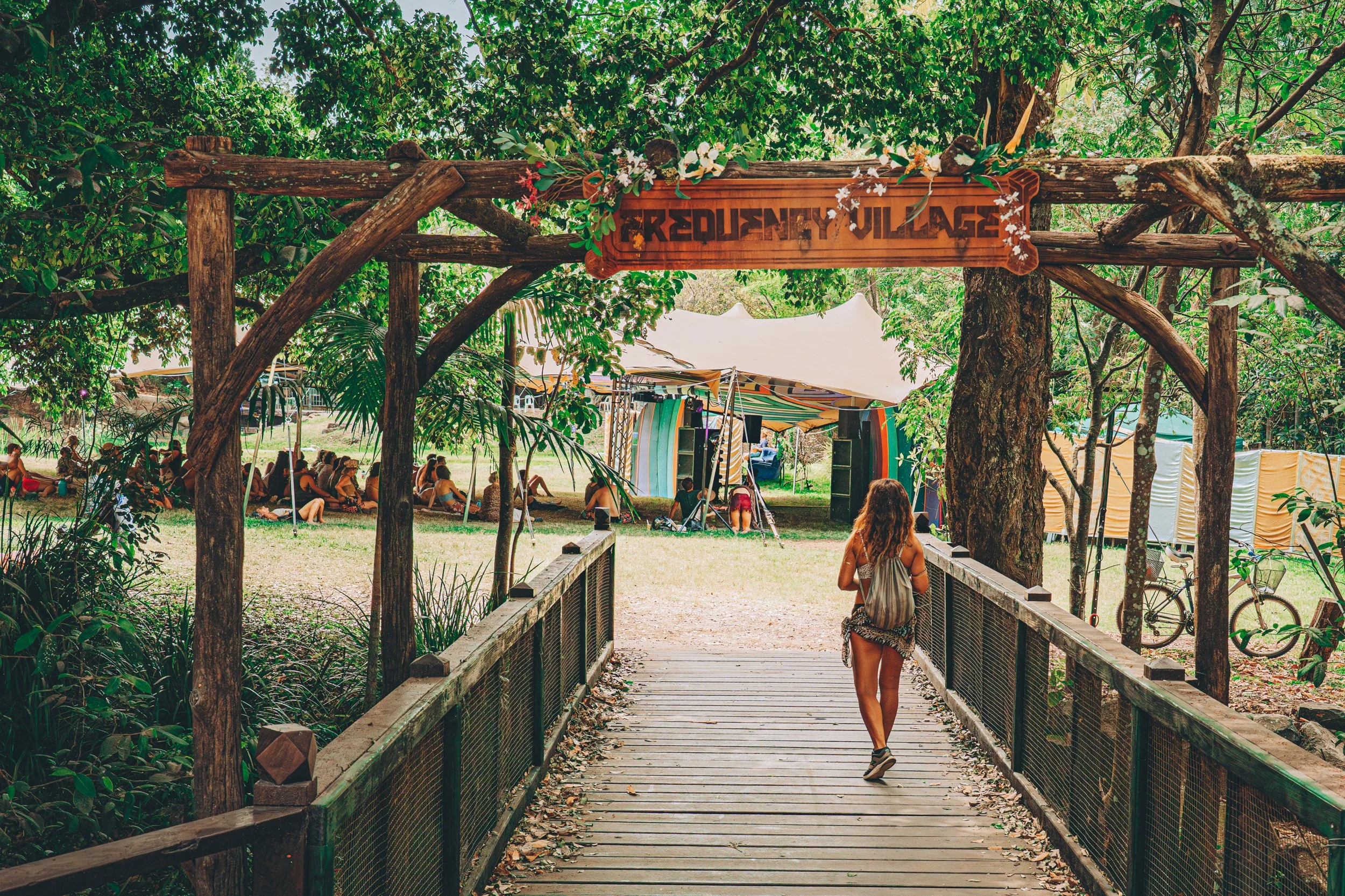 A person with long hair, wearing a backpack and shorts, walks across a wooden bridge toward a festival area called 'Frequency Village' in a wooded outdoor setting. There are tents and people sitting on the grass in the background.