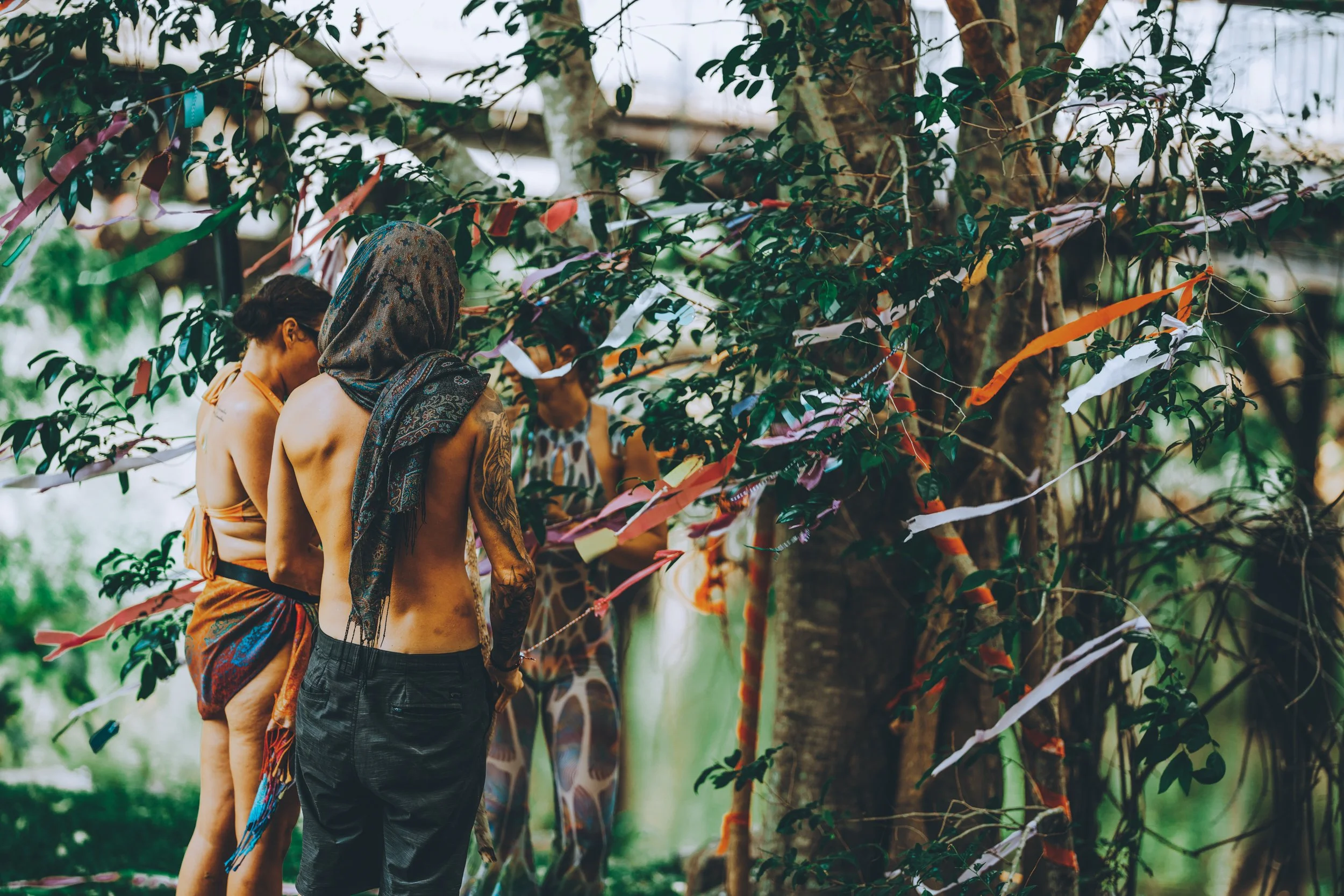 Three people in a garden or forest setting, standing near a tree decorated with colorful ribbons or fabric strips, with lush green foliage around.