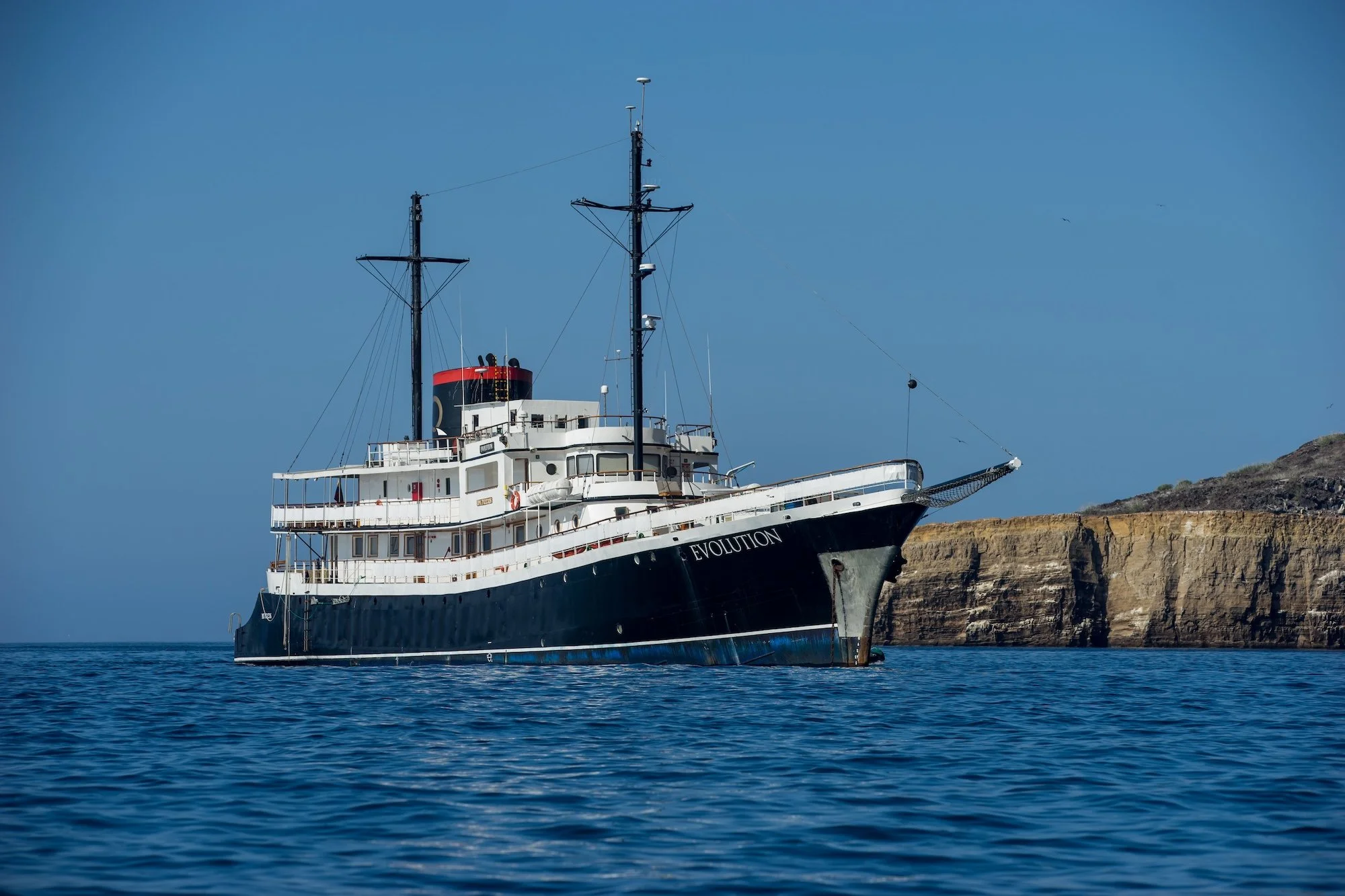 A large vintage ship named EVOLUTION sailing on calm ocean water near a rocky coastal cliff.