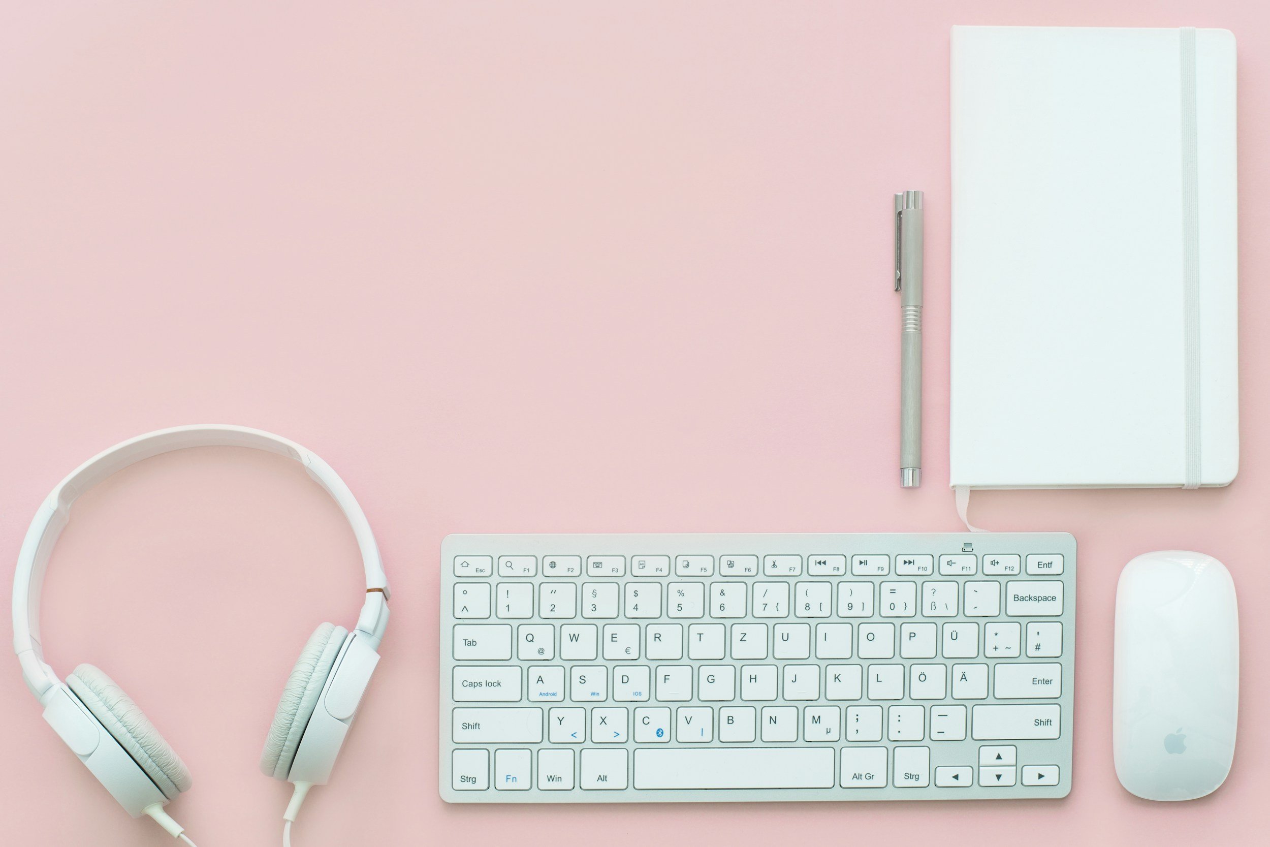 Flat lay of a white computer keyboard, white wireless mouse, white headphones, silver pen, white notebook with an elastic band on a pink background.