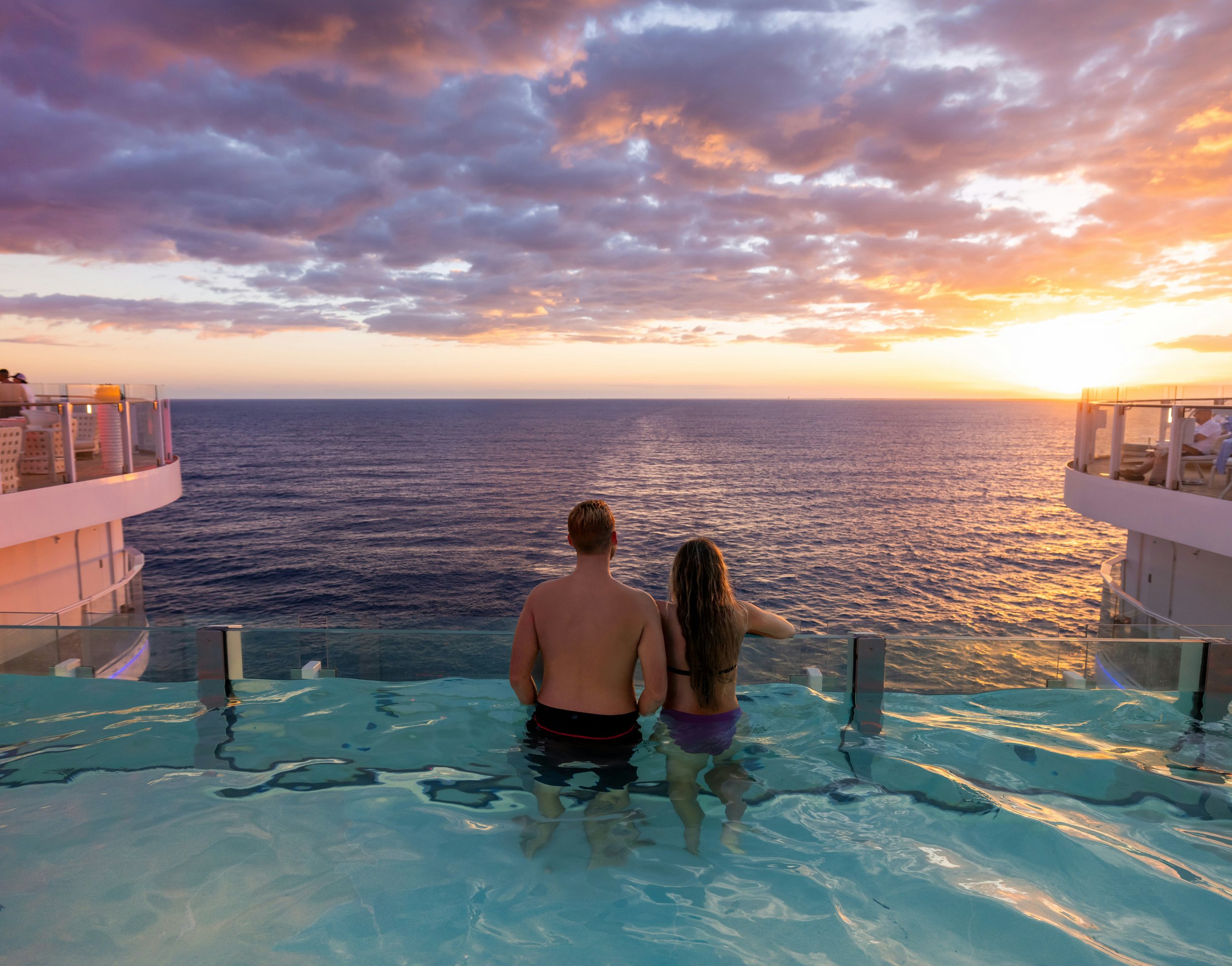 A man and woman in swimsuits standing in an infinity pool on a cruise ship, looking out at the ocean and sunset.