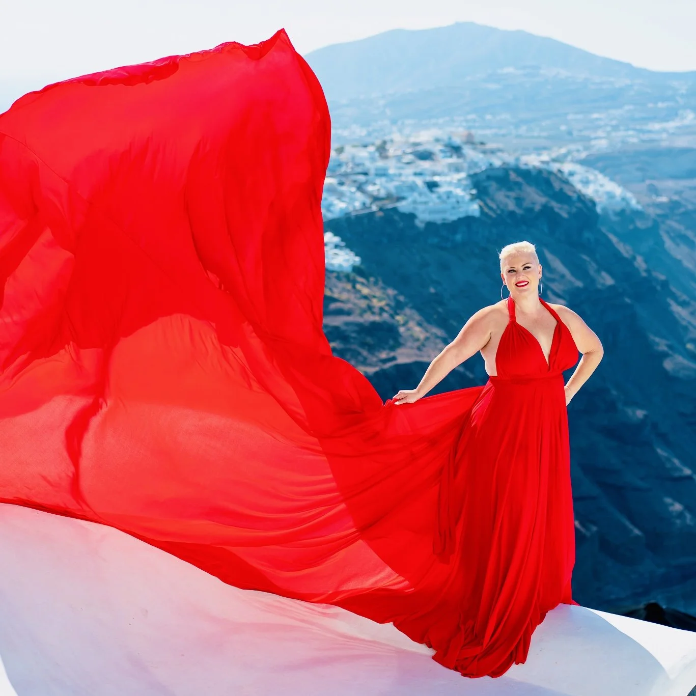 A woman with platinum blonde hair wearing a red dress with a long train stands on a white surface with a mountainous landscape in the background.