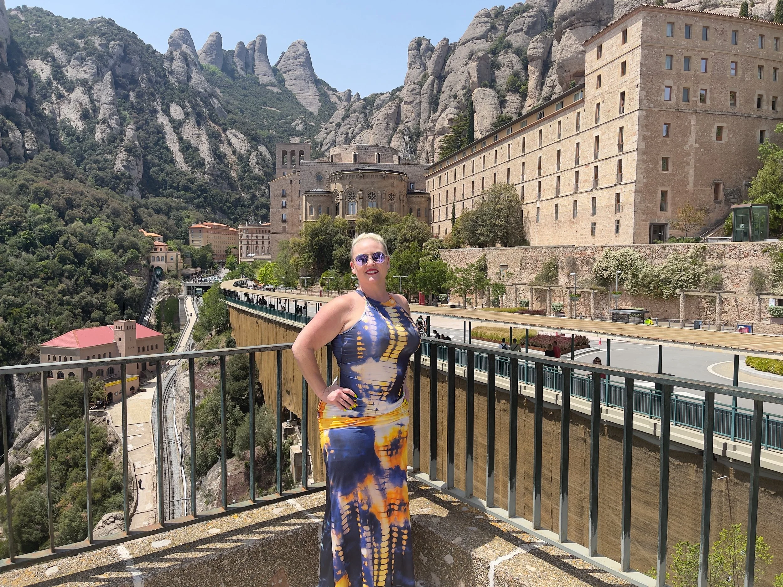 A woman in a colorful tie-dye dress standing on a balcony overlooking Montserrat mountain in Catalonia, Spain, with rocky peaks and historic buildings in the background.