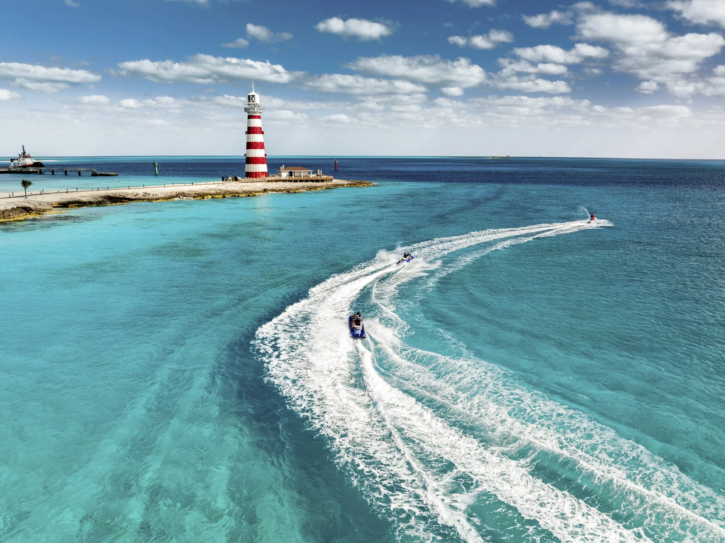Three jet skis riding in turquoise waters near a lighthouse on a coastal area, with a partly cloudy sky overhead.