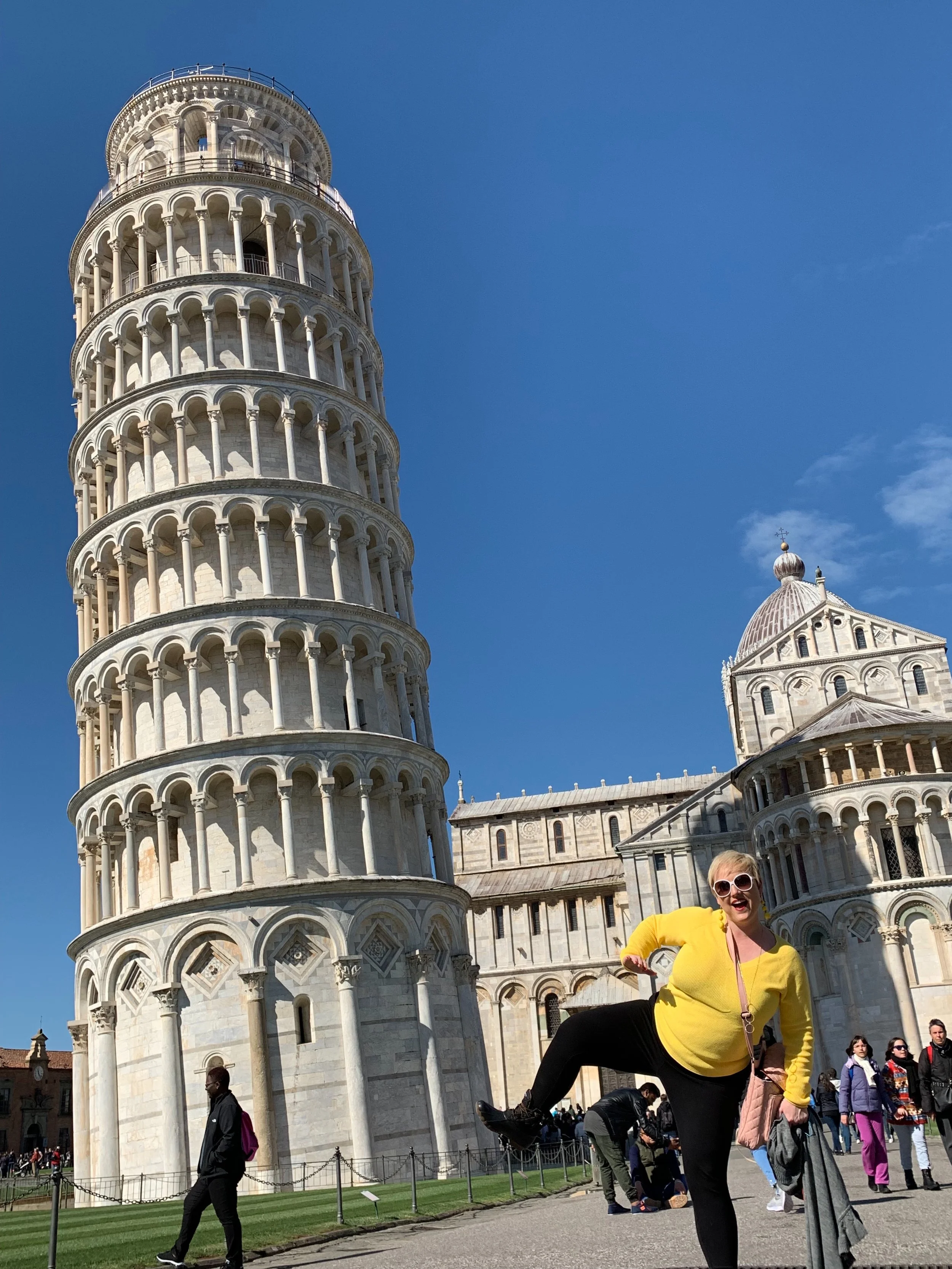A woman in a yellow top and black pants posing in front of the Leaning Tower of Pisa, with tourists and historic buildings in the background under a clear blue sky.