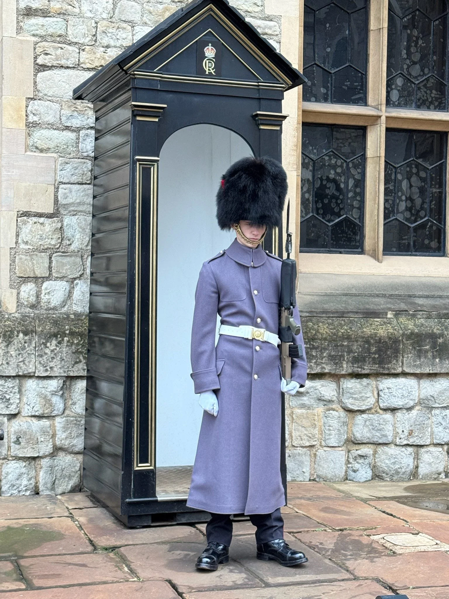A British royal guard standing next to a guardhouse outside a stone building with stained glass windows.