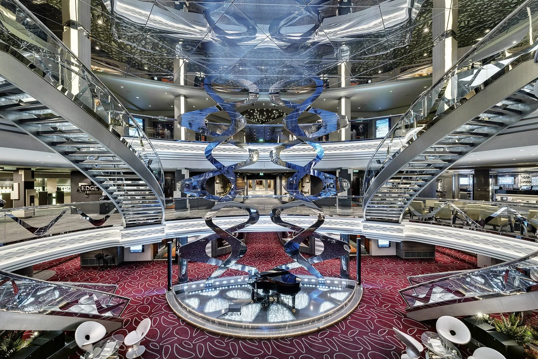 Interior of a modern multi-level shopping mall or hotel with a large Phillips-style spiral staircase, reflective surfaces, a grand piano on a circular platform, red patterned carpet, and seating areas.