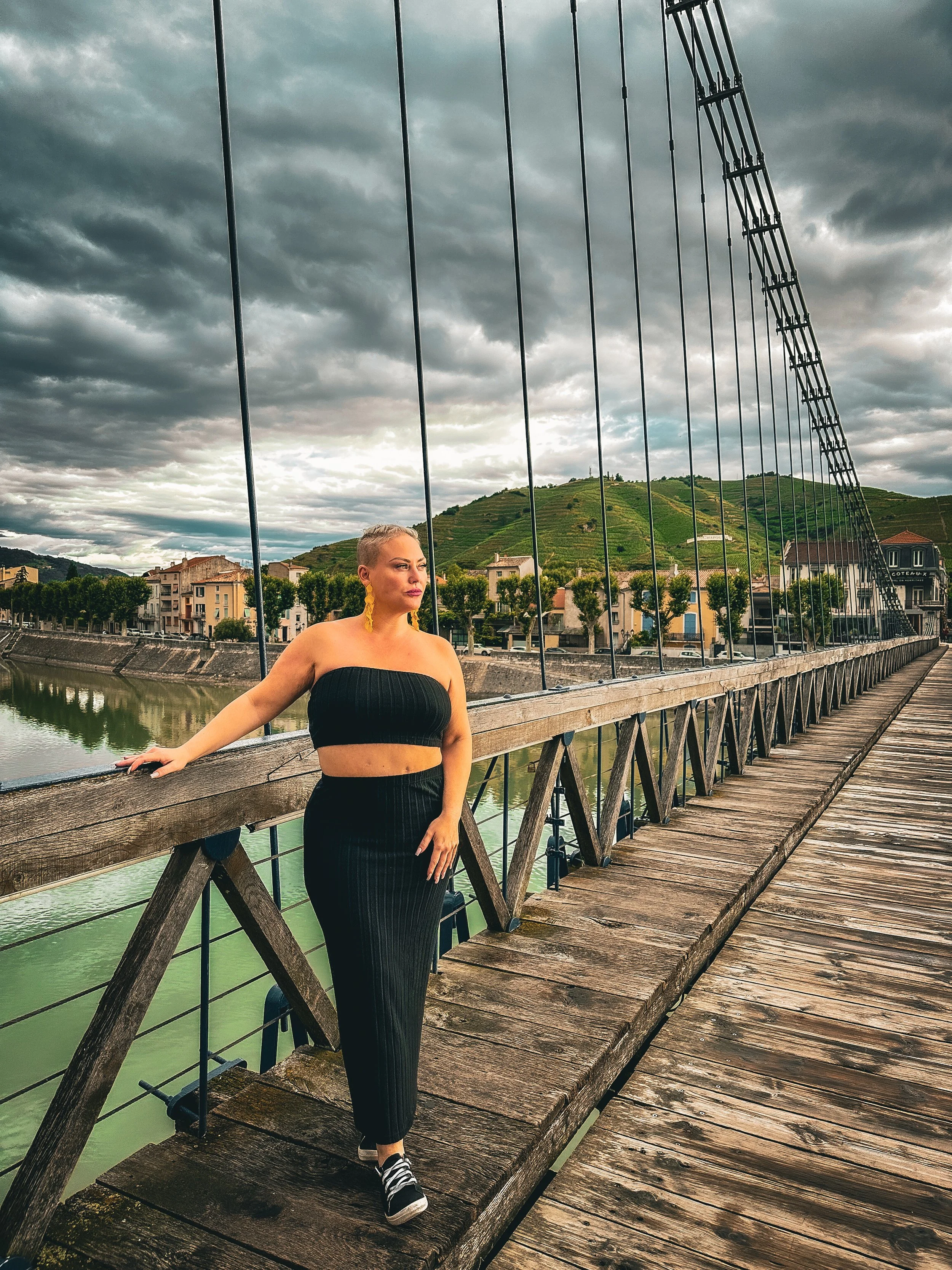 A woman in a black strapless top and high-waisted black pants posing on a wooden bridge over a river with a hilly landscape and cloudy sky in the background.