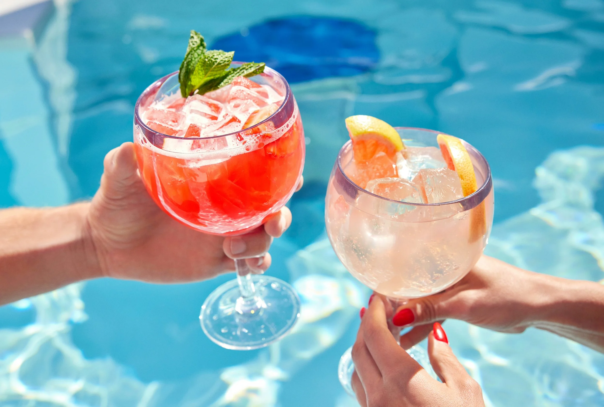 Two women with red painted nails holding large stemmed glasses filled with pinkish-red and clear cocktails with lemon slices and mint, near a pool with blue water.