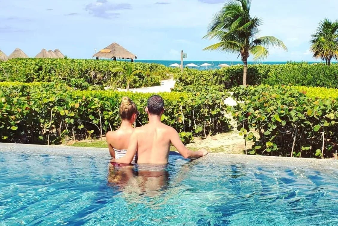 A couple in a pool facing a tropical beach landscape with palm trees and thatched huts.