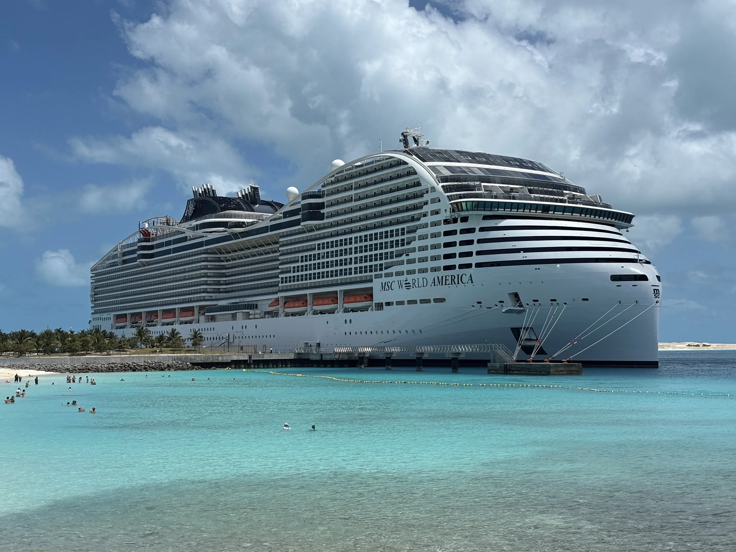Large cruise ship docked at a tropical beach with turquoise water, palm trees, and people swimming and relaxing in the water.
