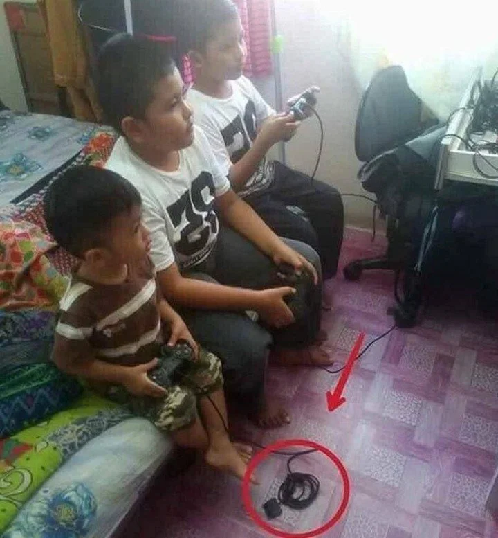 three boys sit on a bed holding video game controllers. the youngest boy's controller is obviously unplugged, circled with a red arrow pointing to it