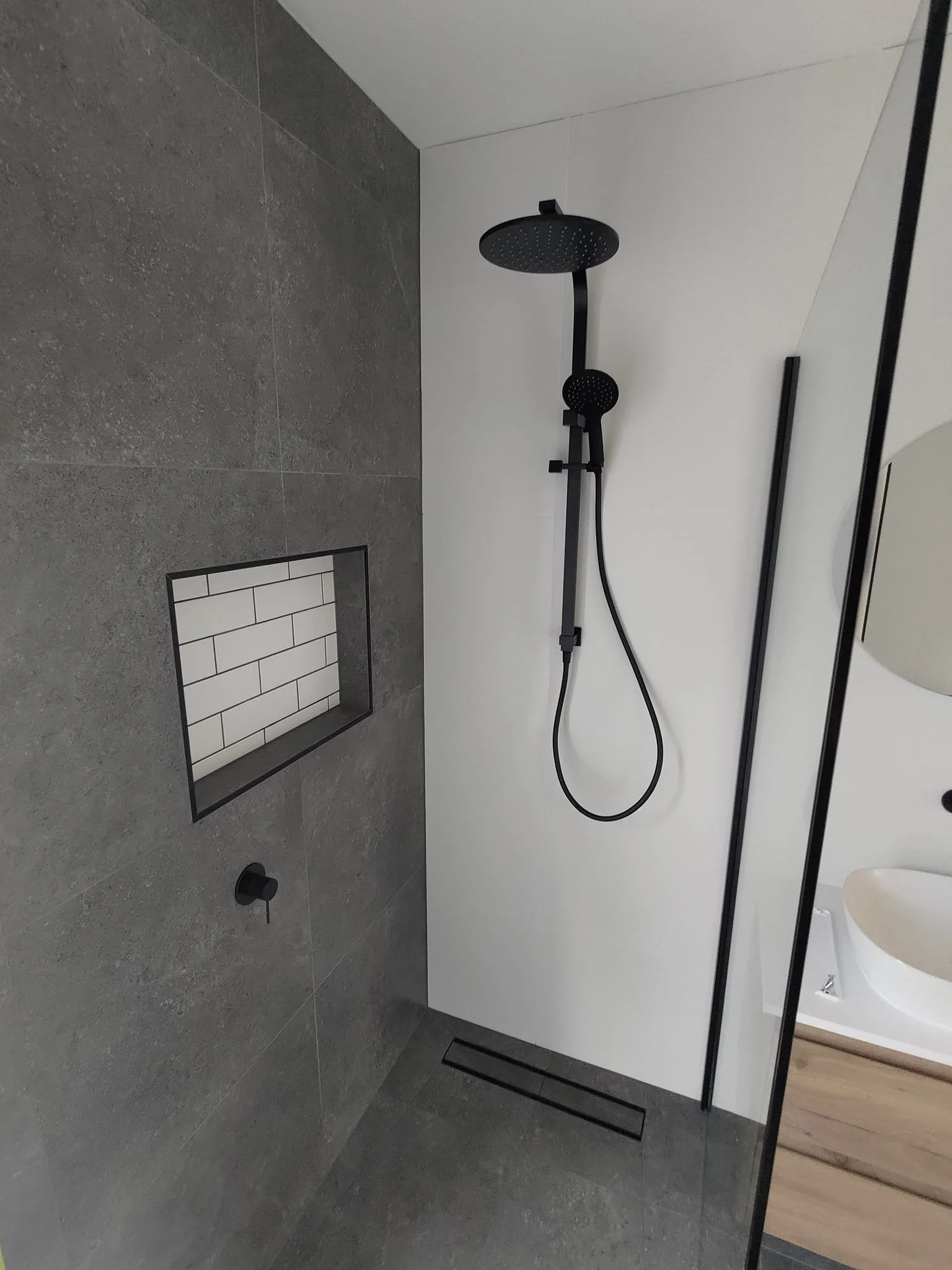 Modern bathroom with dark gray tiled shower, black shower fixtures, a niche with white subway tiles, and a wooden vanity with a white sink.