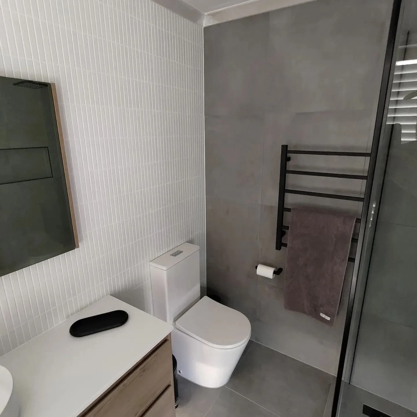 Modern bathroom with gray tiles, a toilet, a black towel rack, and a wooden vanity with a black soap dish.