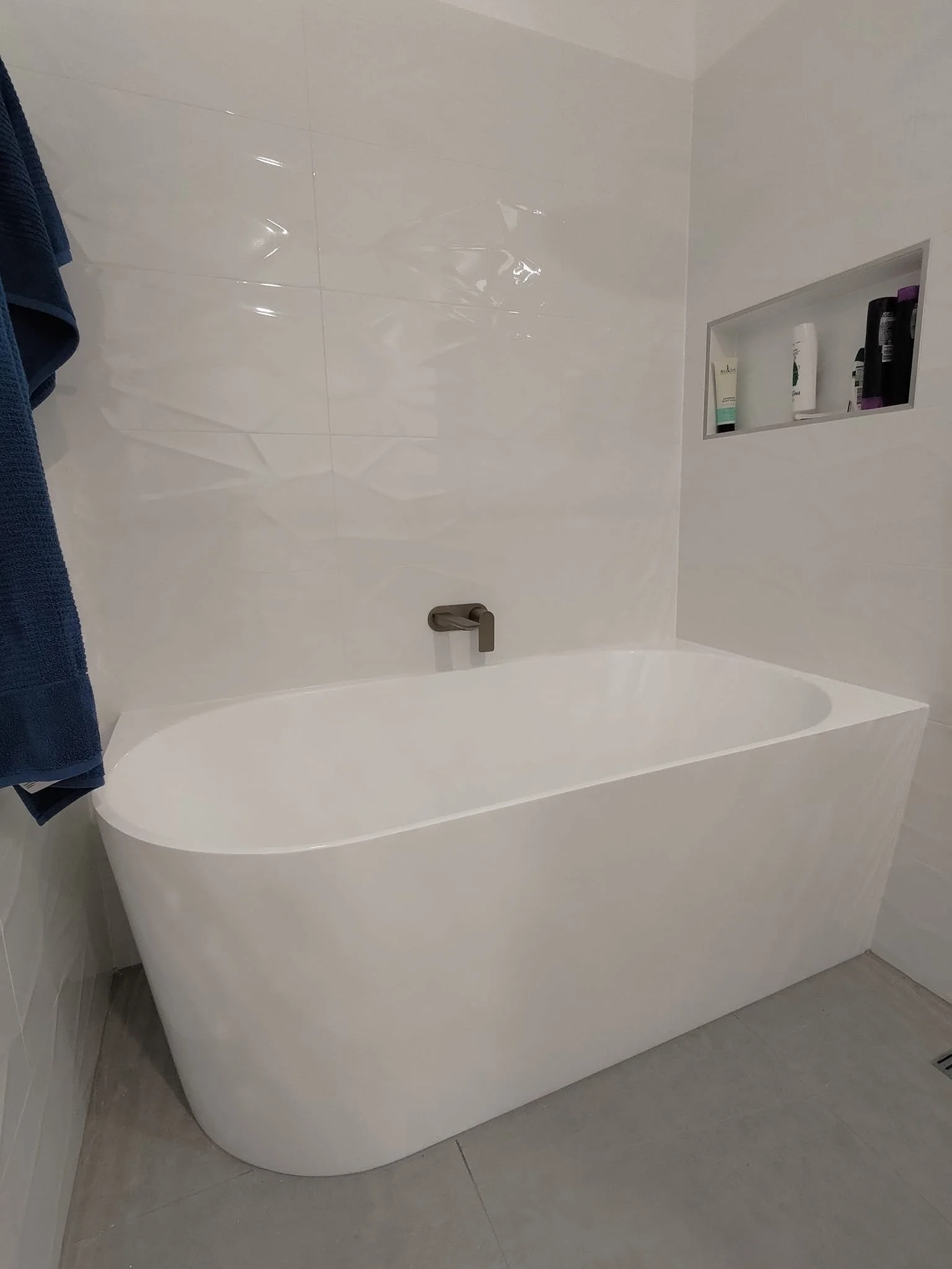 Modern white bathtub with a wall-mounted faucet, blue towel hanging, and built-in shelf with toiletries.