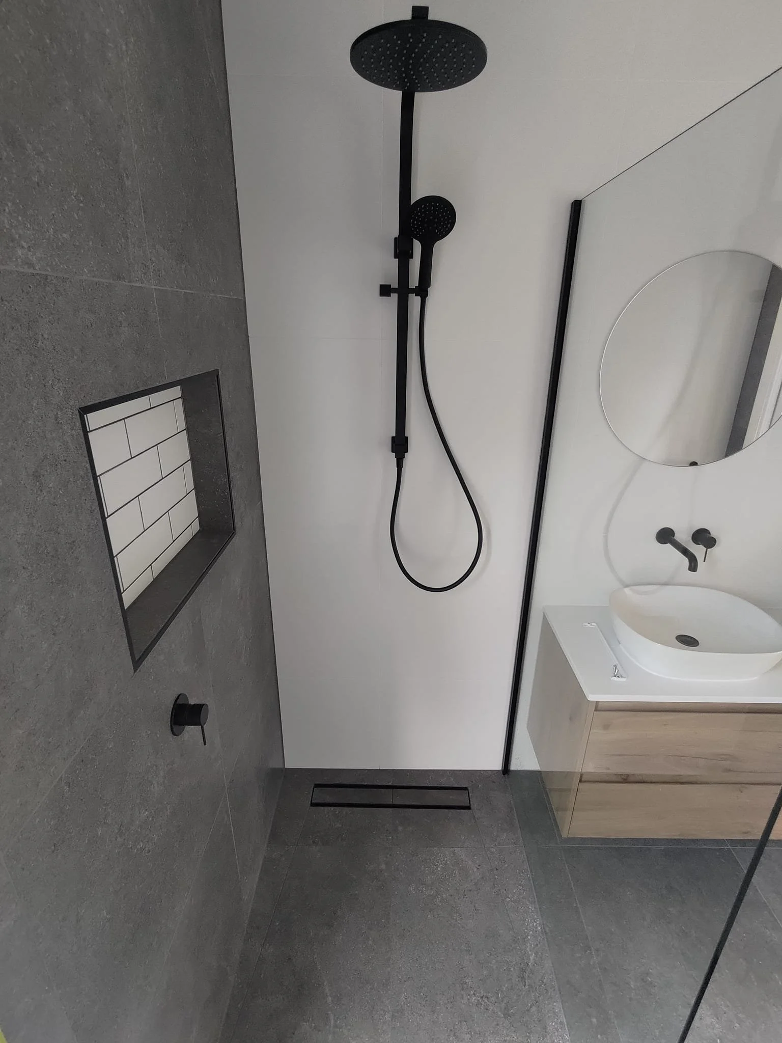 Modern bathroom with gray tiles, walk-in shower, overhead and hand-held black showerheads, wall niche with white subway tiles, floating wooden vanity, round mirror, and white basin sink.