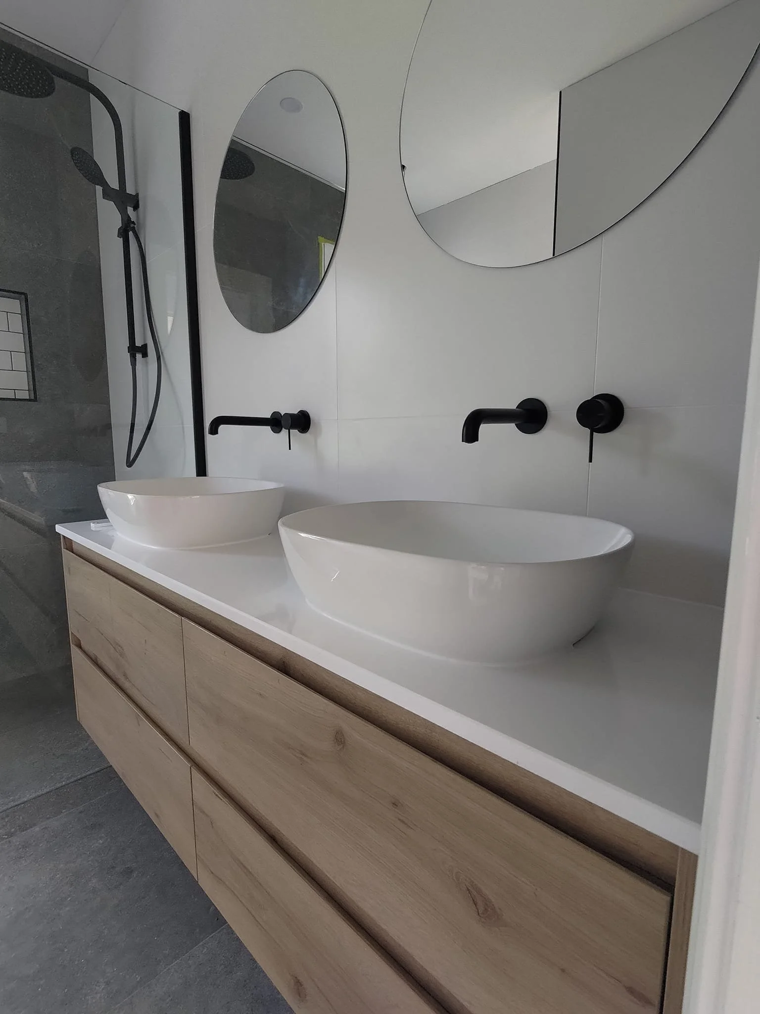 Modern bathroom with two round mirrors, two white vessel sinks on a wooden vanity, and matte black faucets.