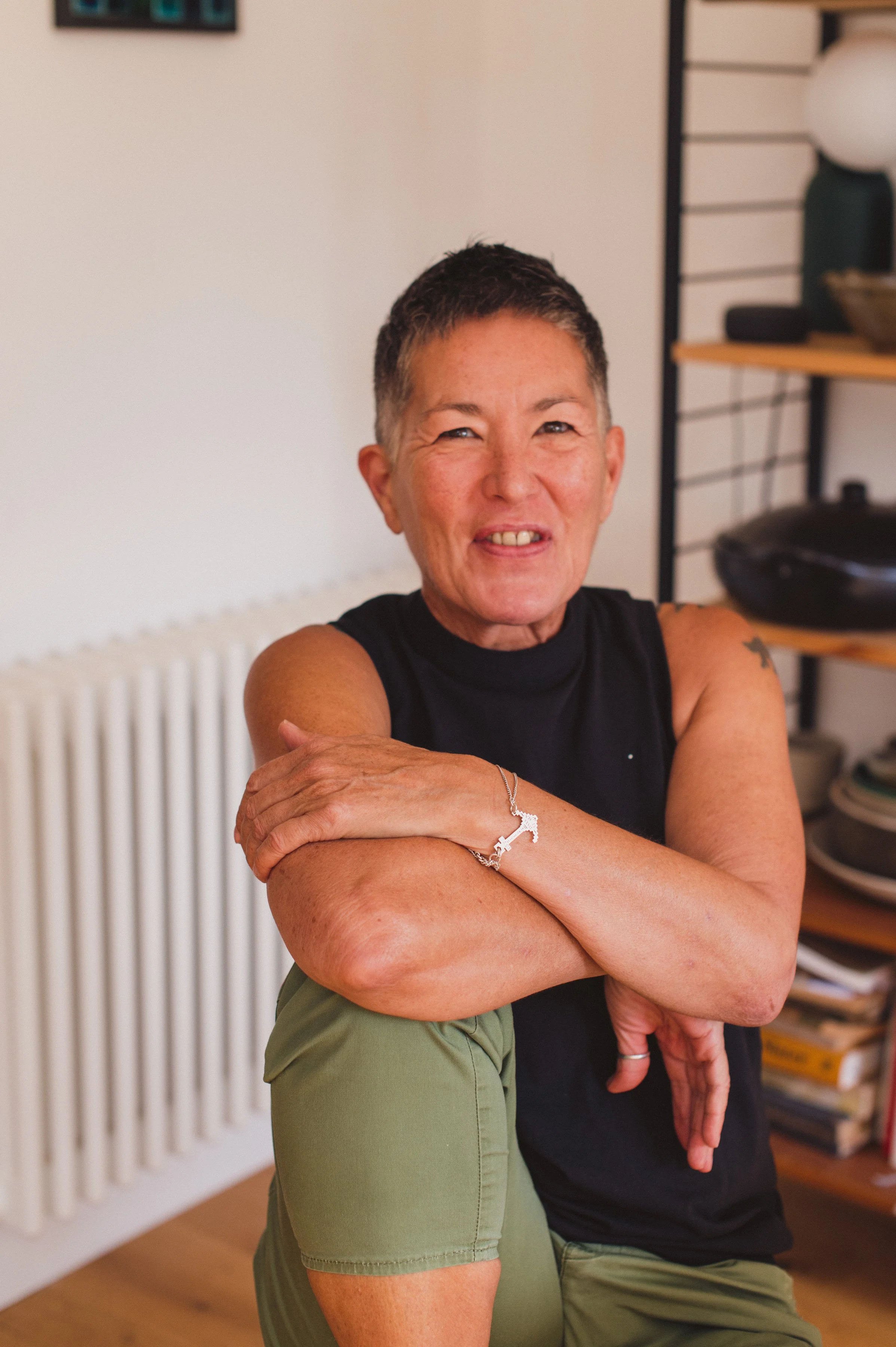A woman with short hair, wearing a sleeveless black shirt, smiling and sitting indoors, with her arms crossed on her knee.