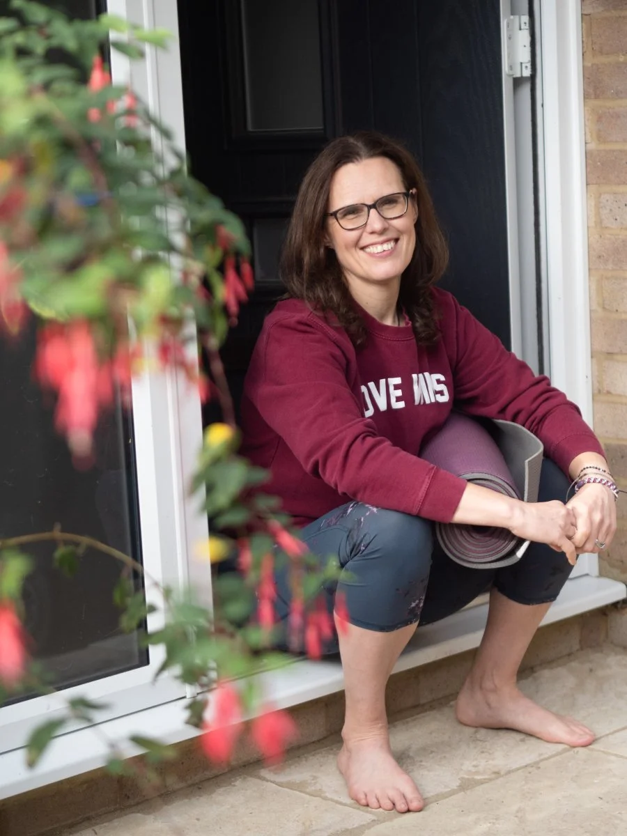 A smiling woman with glasses, wearing a maroon sweatshirt and dark pants, sitting barefoot on a porch step, holding a yoga mat, with flowers in the foreground and a black door behind her.