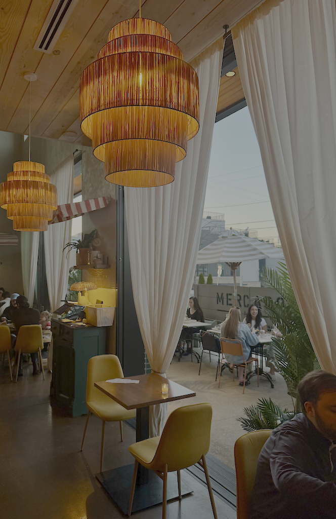 Interior of a restaurant or cafe with warm lighting, large hanging lamps, cream curtains, tables with yellow and orange chairs, and people dining outdoors under umbrellas.