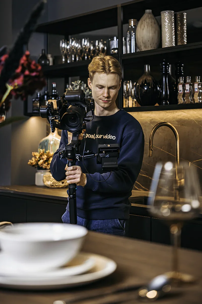 A person with red hair operating a professional video camera in a modern kitchen or dining area. There are glasses, bottles, and decorative vases on shelves behind them, with a table set with dishes and a wine glass in the foreground.