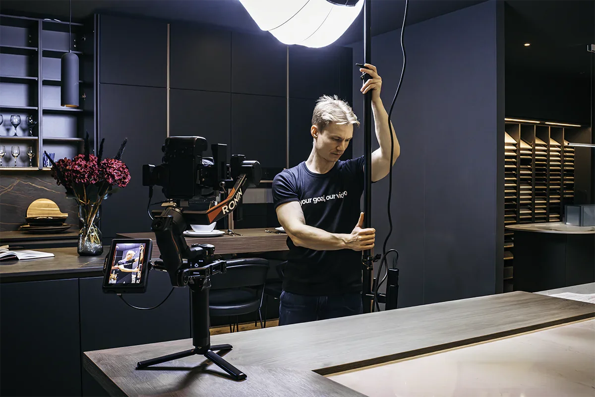 A person in a black t-shirt adjusts a large overhead light in a modern kitchen or studio setting, with a camera on a stabilizer on the counter.