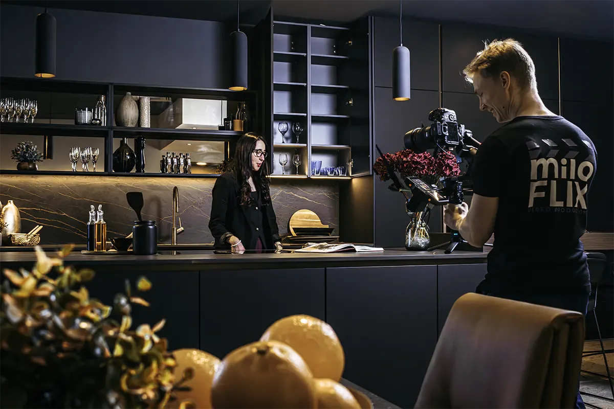 A woman is being filmed by a man in a modern kitchen with black cabinets and gold accents, while a camera operator films her holding an open book.