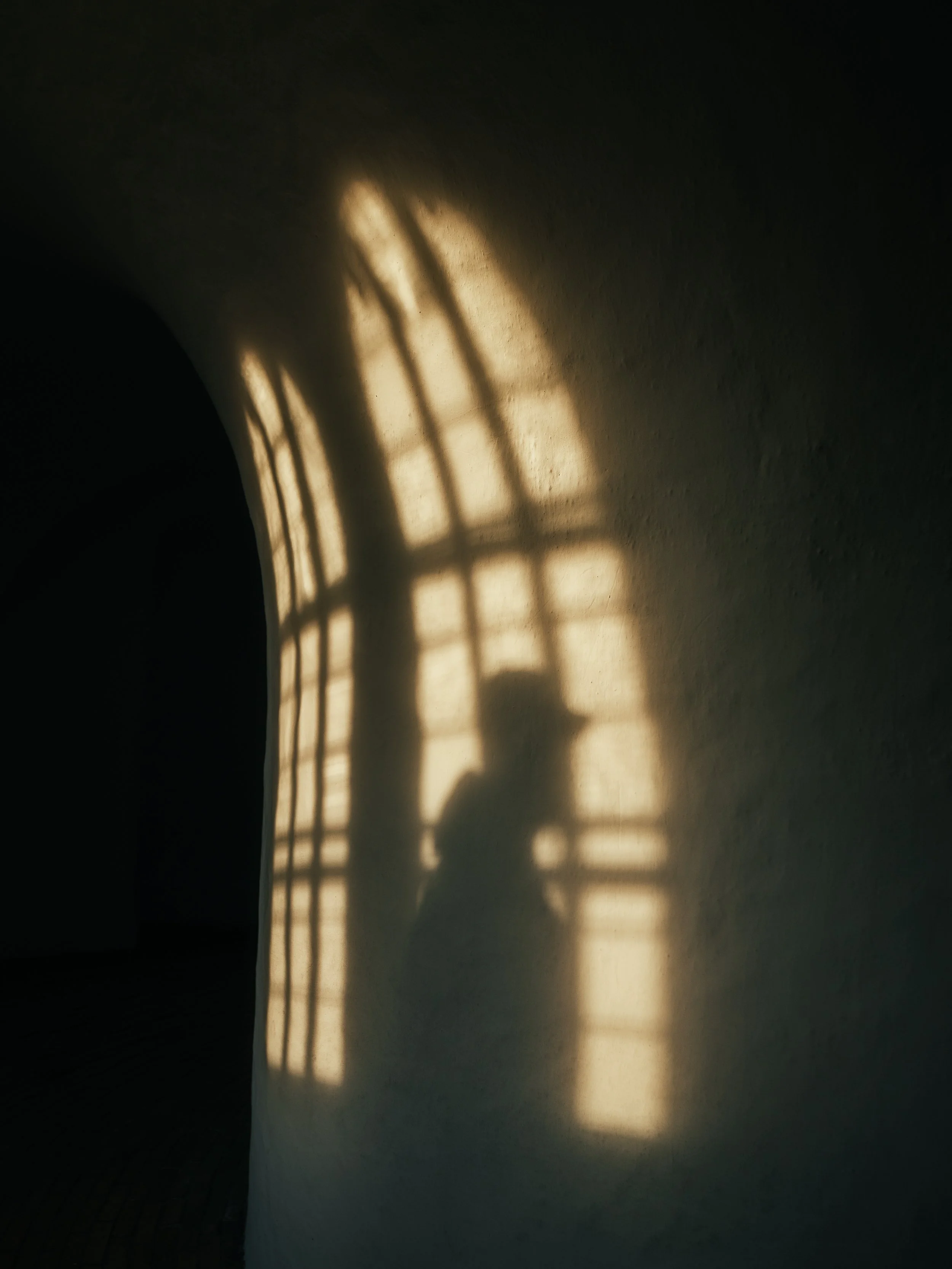 The shadow of a person wears a hat and leans against a textured wall near a window with grid-patterned sunlight.