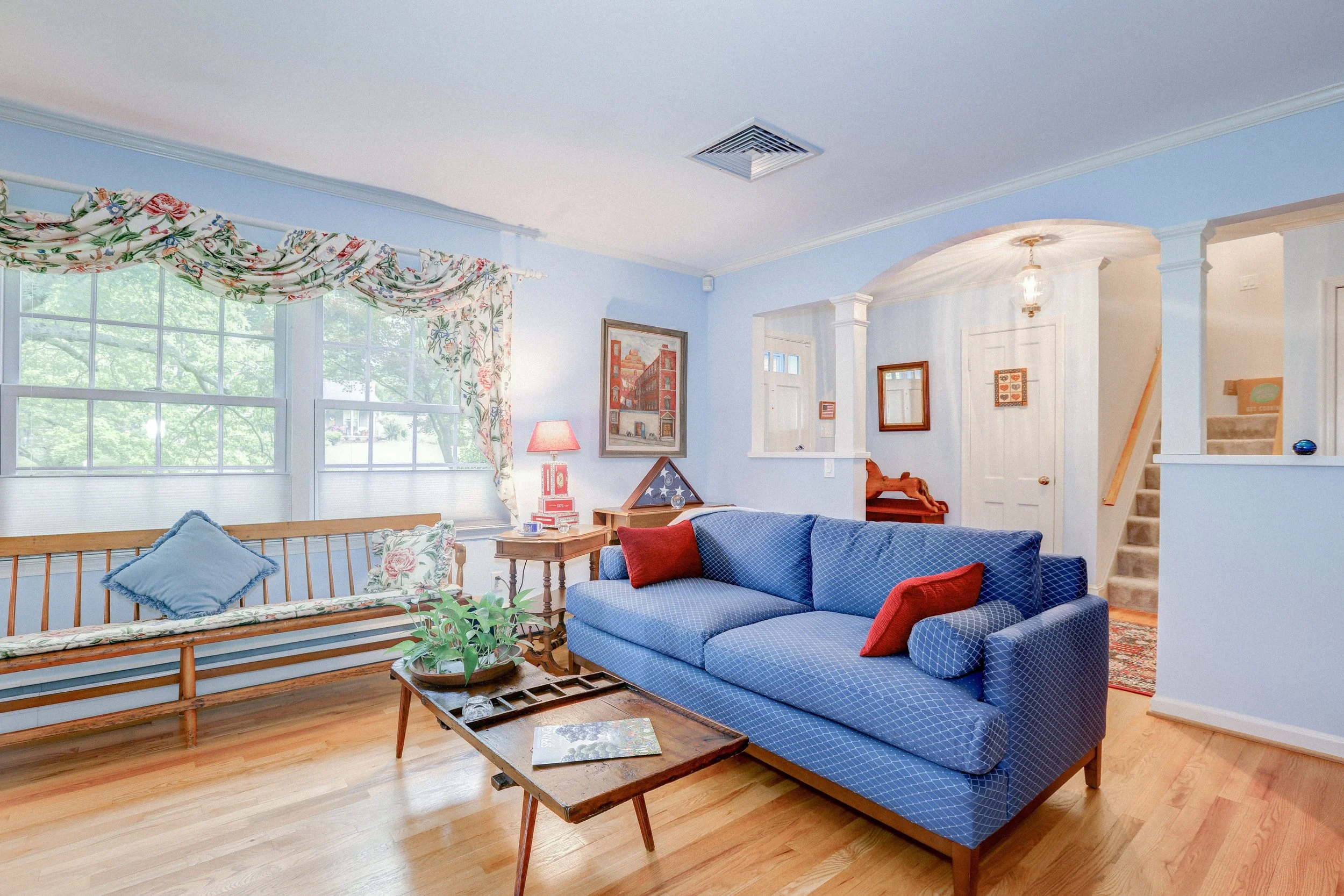 Cozy living room with blue couch, red accents, floral curtains, wooden bench, and coffee table with plants. Light blue walls and natural light from large windows.