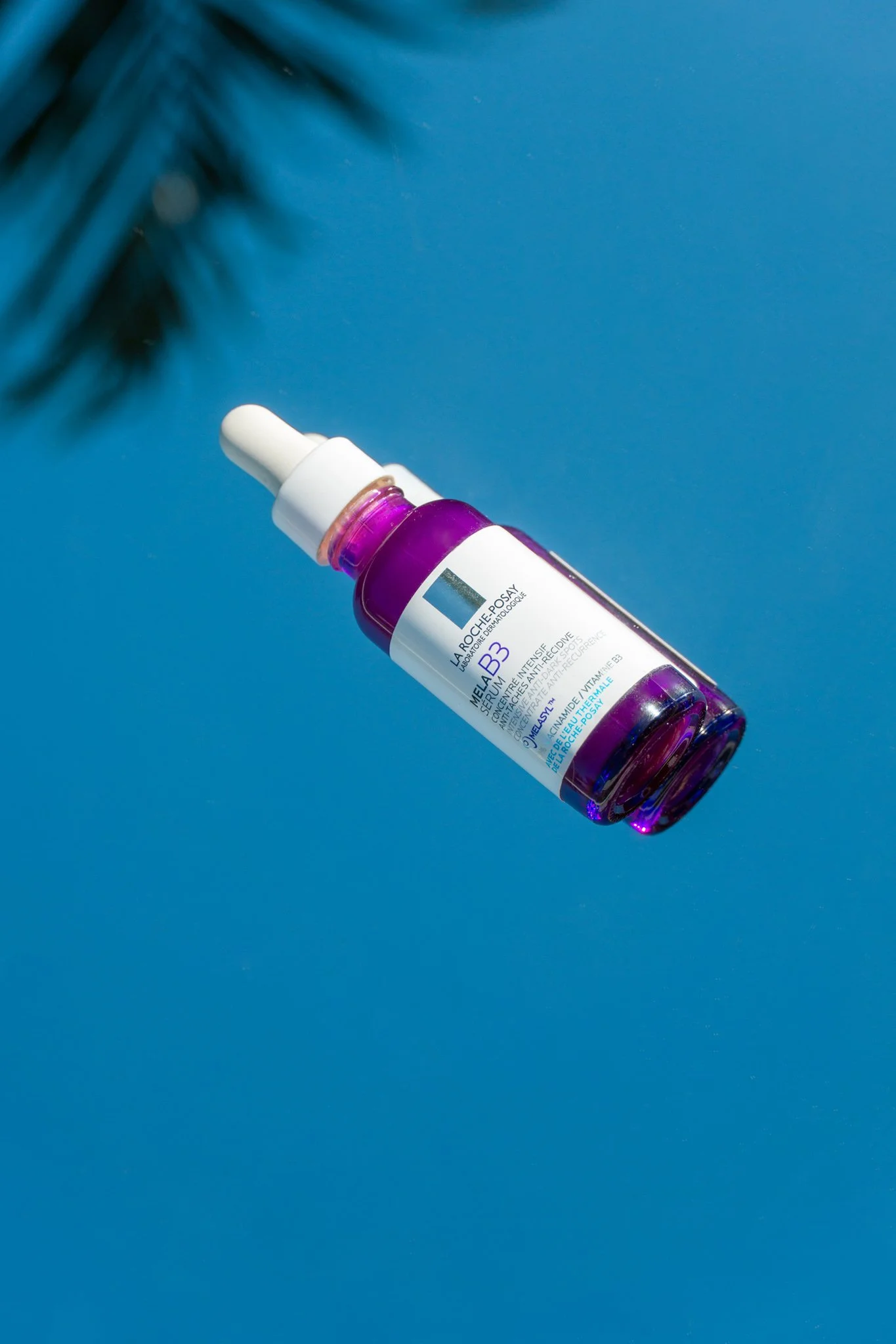 A skin serum bottle with a dropper cap is placed on a blue background, with a palm frond shadow in the upper left corner.
