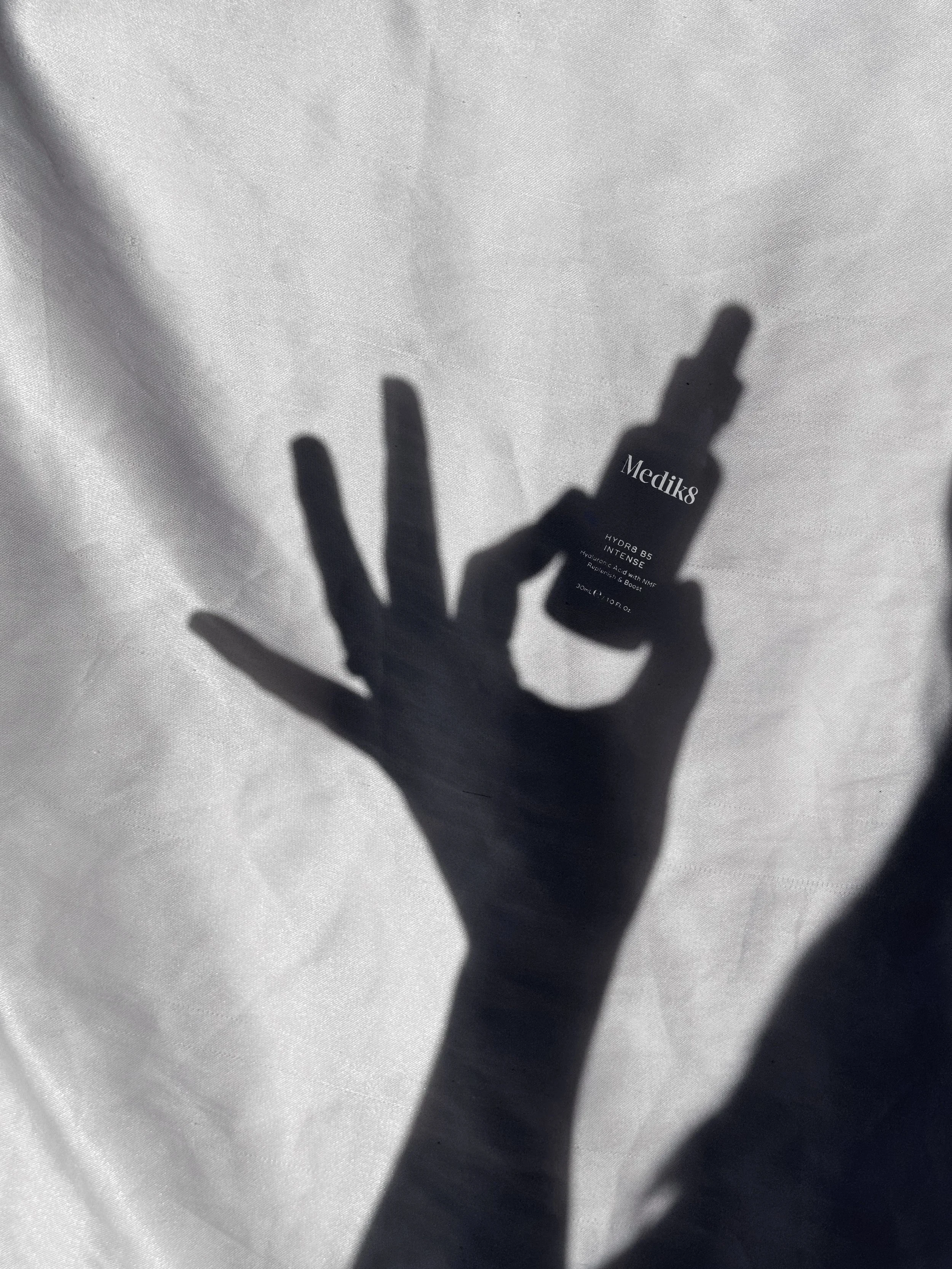 A shadow of a hand holding a dropper bottle of Medik8 skincare product against a white background.