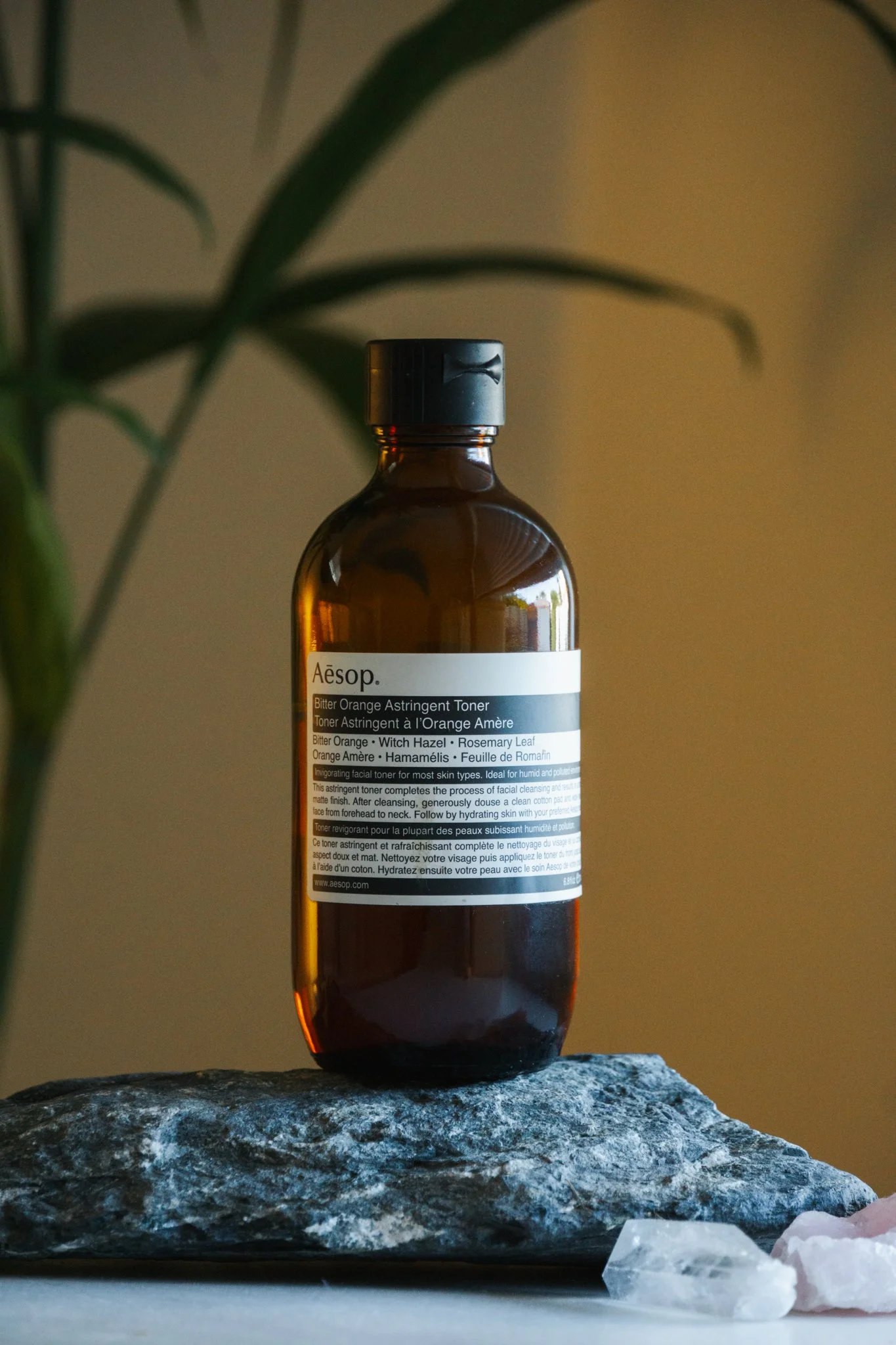 A brown glass bottle of Aesop Bitter Orange Astringent Toner placed on a stone surface with some quartz stones nearby, with a blurred plant in the background.