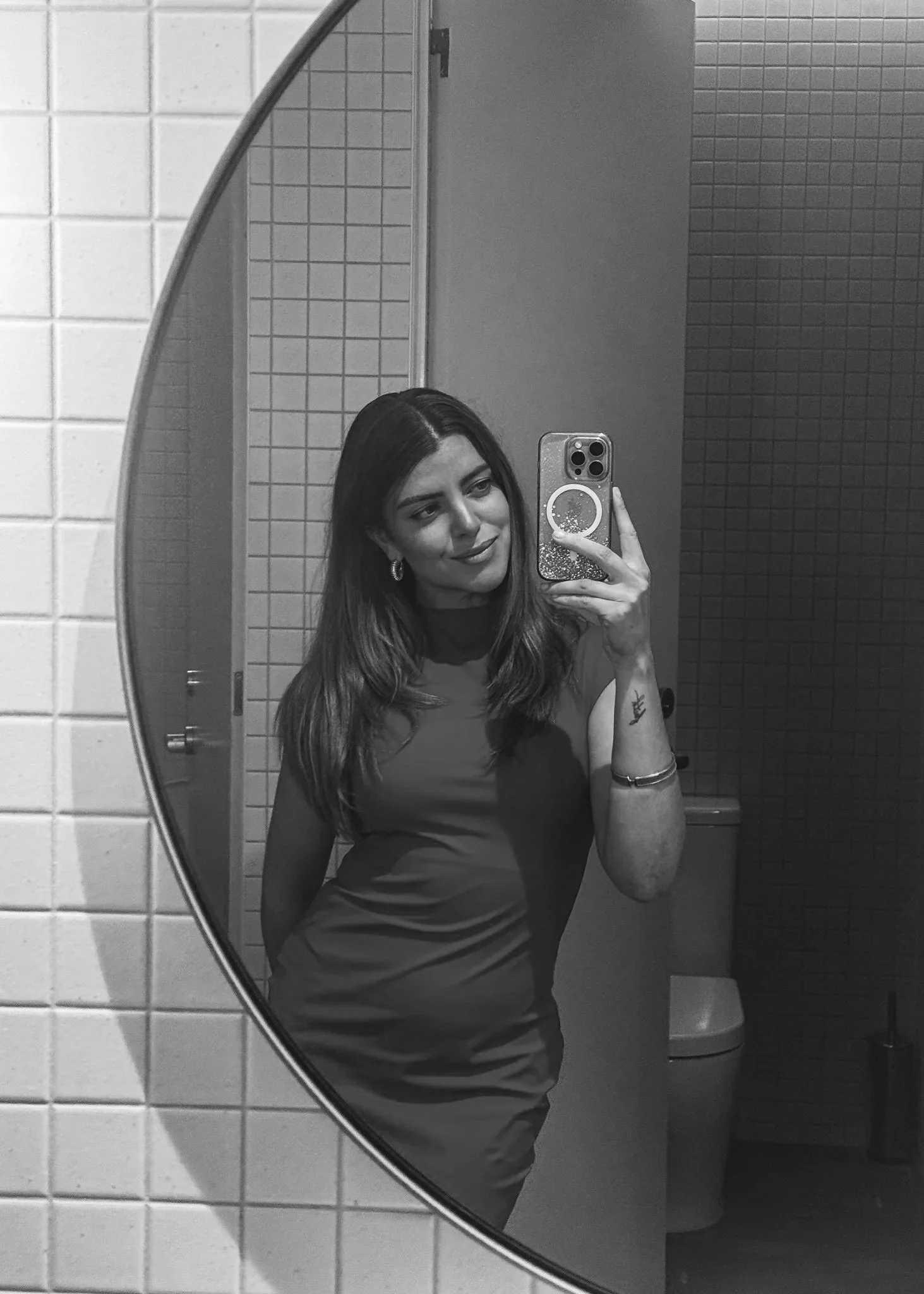 A woman takes a mirror selfie in a public restroom, smiling slightly, with her dark hair down, wearing earrings and a sleeveless dress. The mirror is framed in a rounded shape, and there are tiled walls and a toilet in the background.