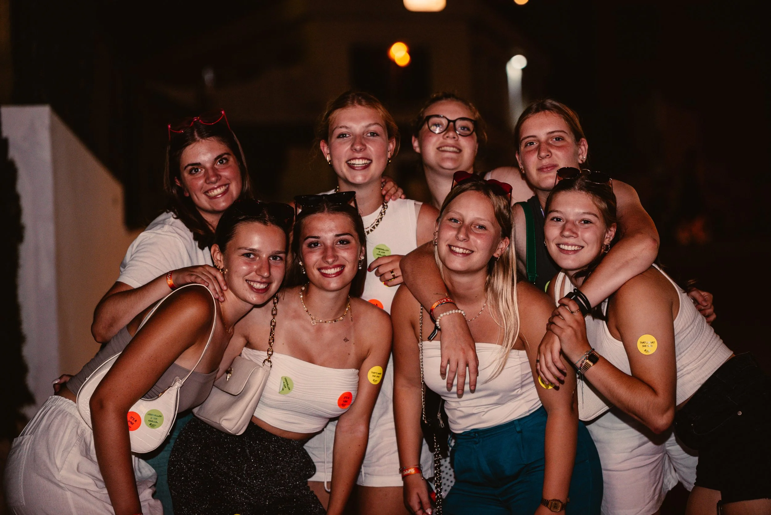Group of young people smiling and posing for a photo at night, wearing casual summer attire and sunglasses, with stickers on their clothes, in an outdoor setting.