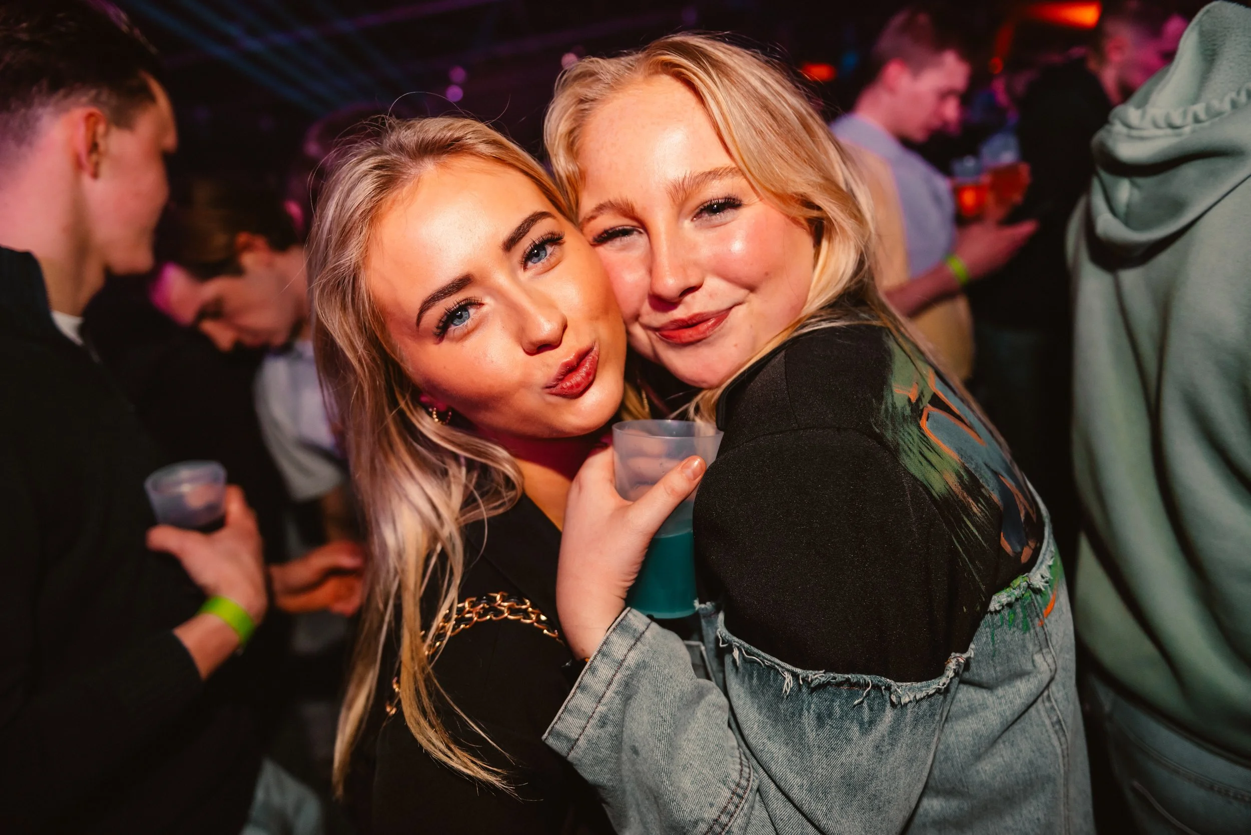 Two women smiling and posing at a party, holding drinks, with people in the background.