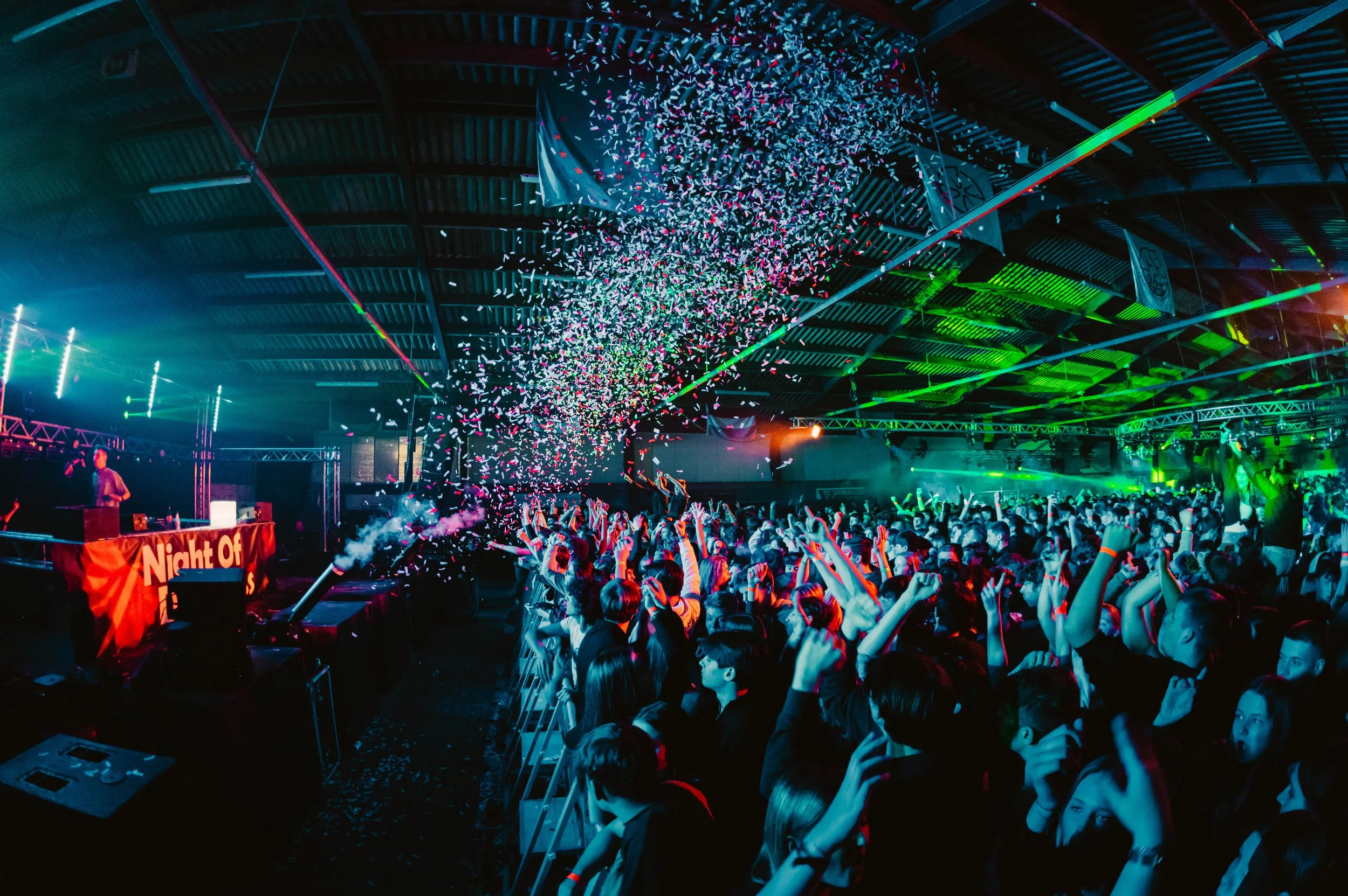 A lively indoor concert scene with a large crowd dancing and cheering. Colorful lights illuminate the venue while confetti falls from above, creating a festive atmosphere. A DJ is performing on stage with equipment in front of them.