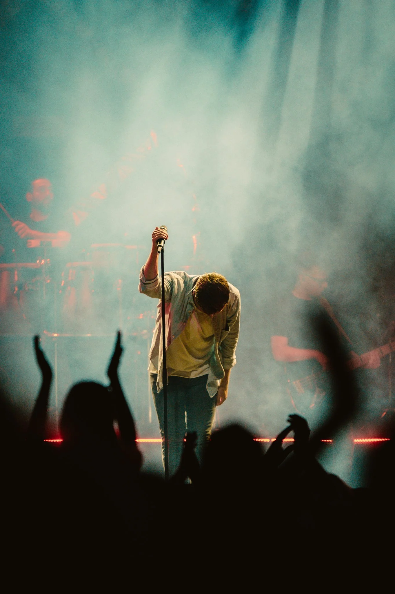 Singer bowing on stage during a concert with a microphone stand, while a drummer plays in the background and the audience claps.
